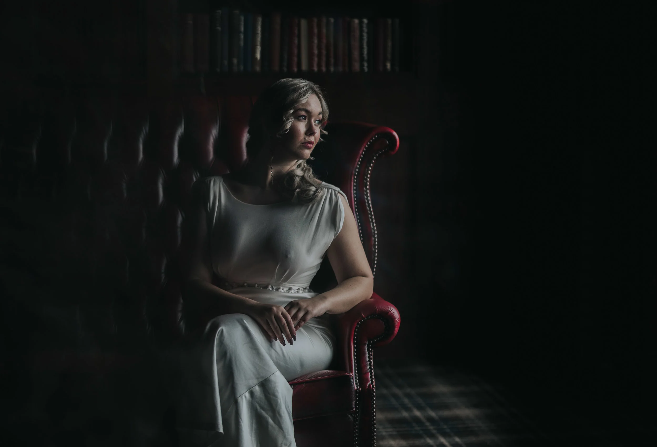 Bride sits on a dark red leather chair looking out of the window with the light flooding her face. The rest of the room is dark.