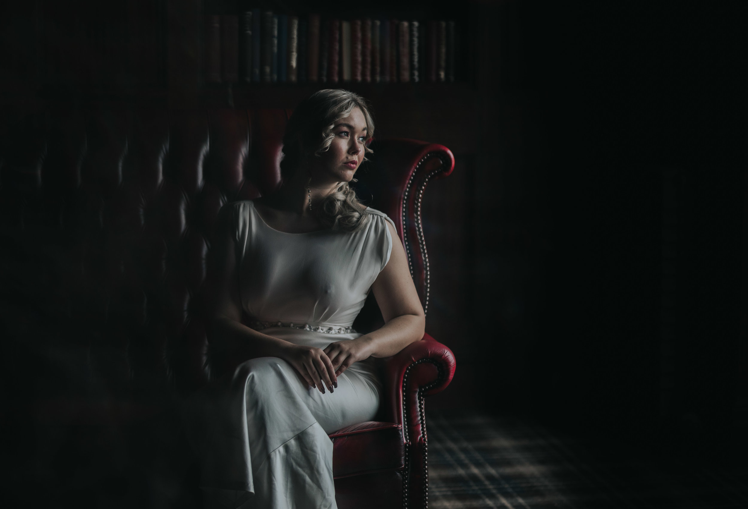Bride sits on a dark red leather chair looking out of the window with the light flooding her face. The rest of the room is dark.