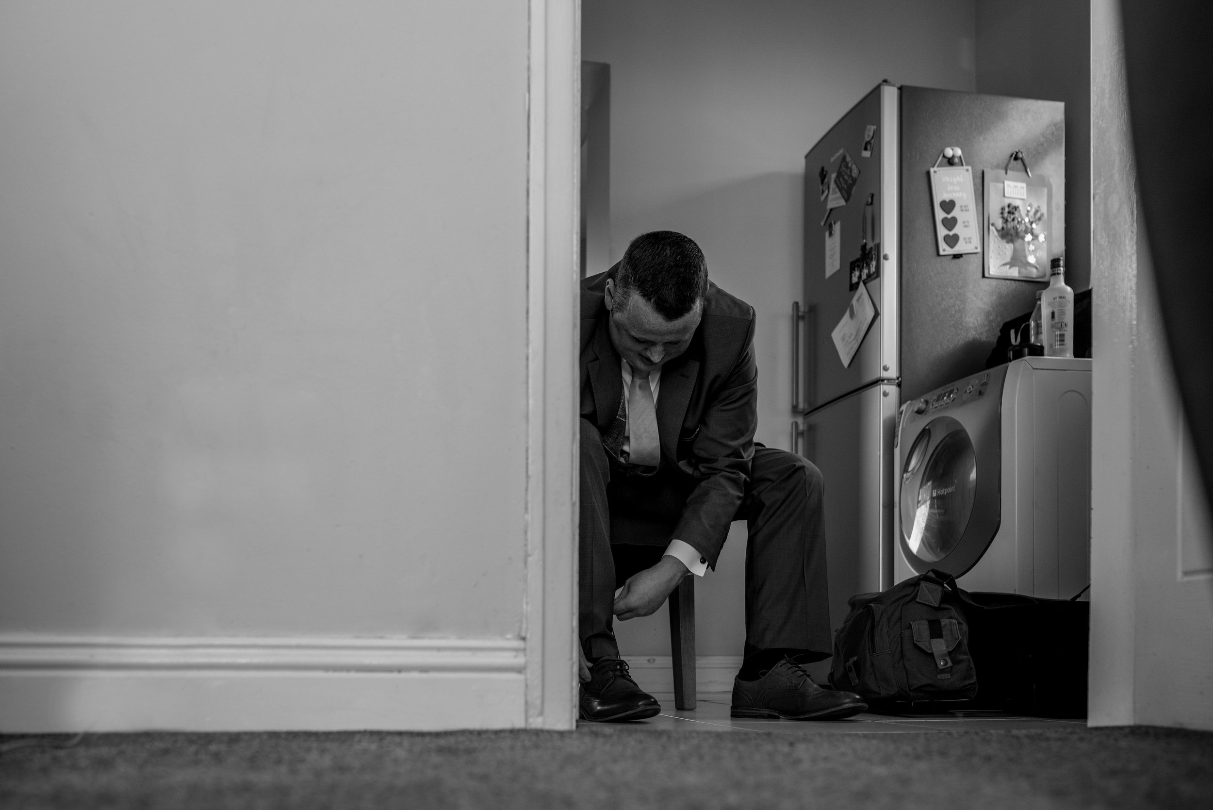 The groom sits on a chair in the kitchen to put his wedding shoes on.