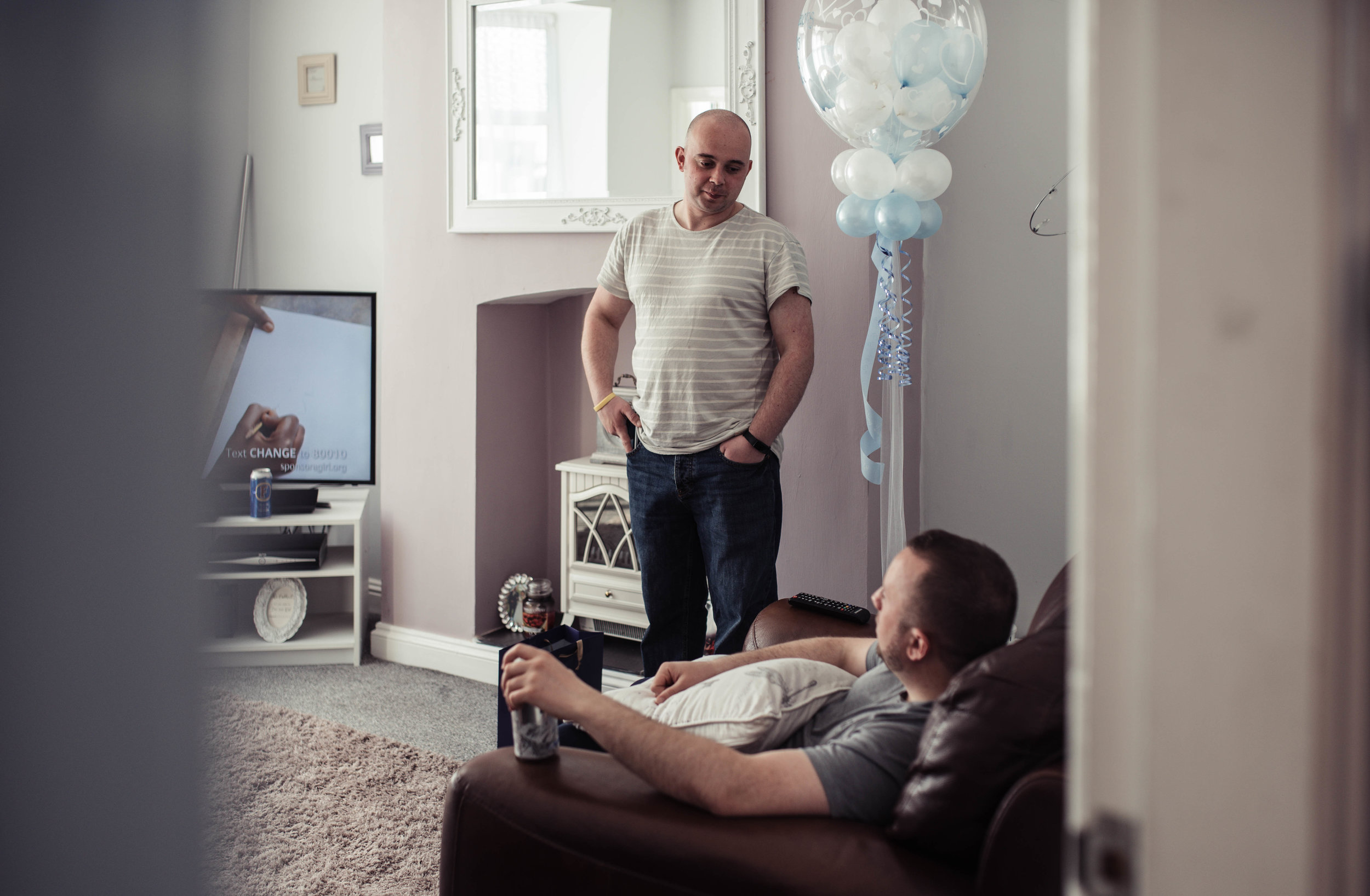 Two groomsmen, one sitting on a chair and another one standing chat to each other in the living room