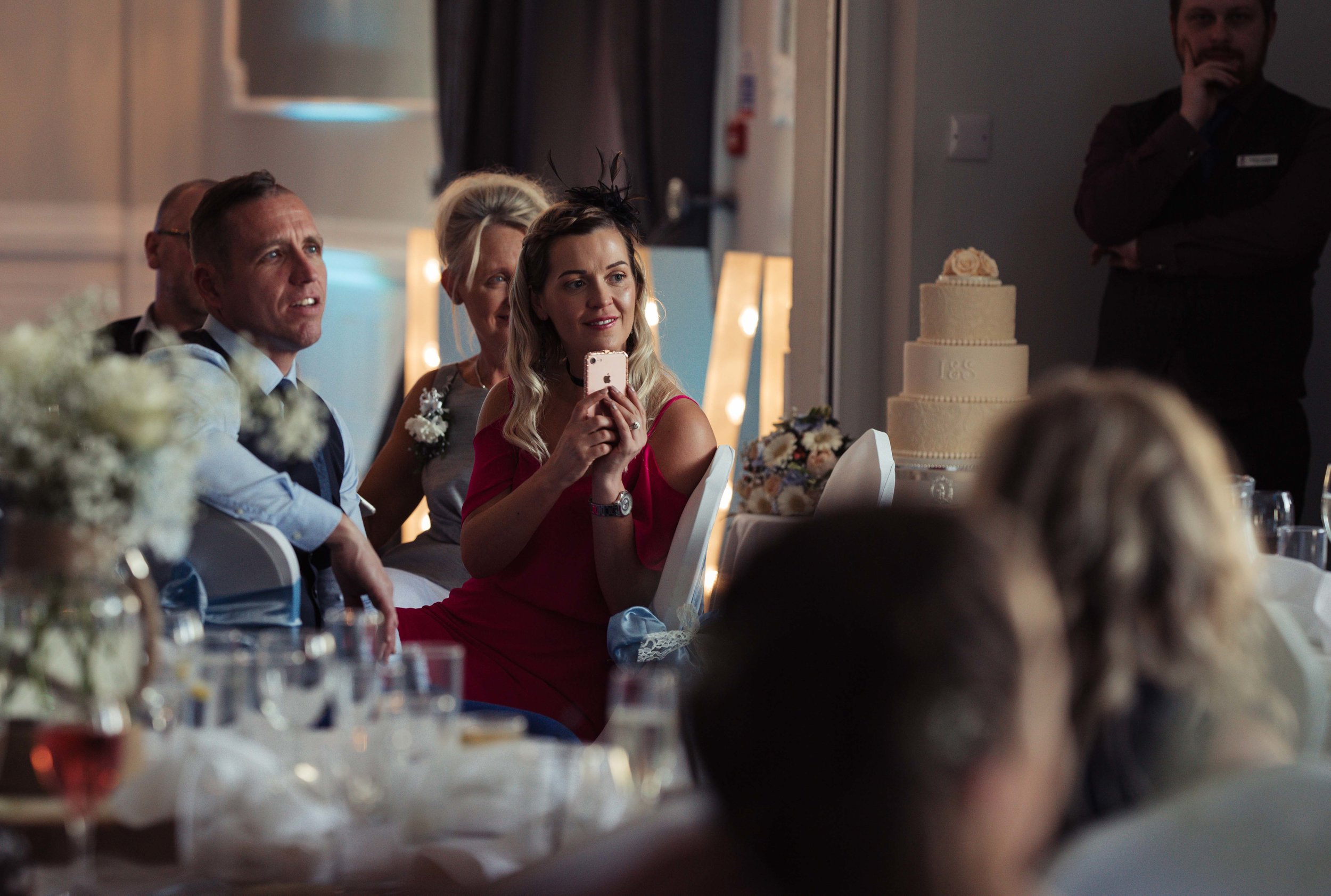 Wedding guest sitting besides the cake sits and films the speeches on her phone