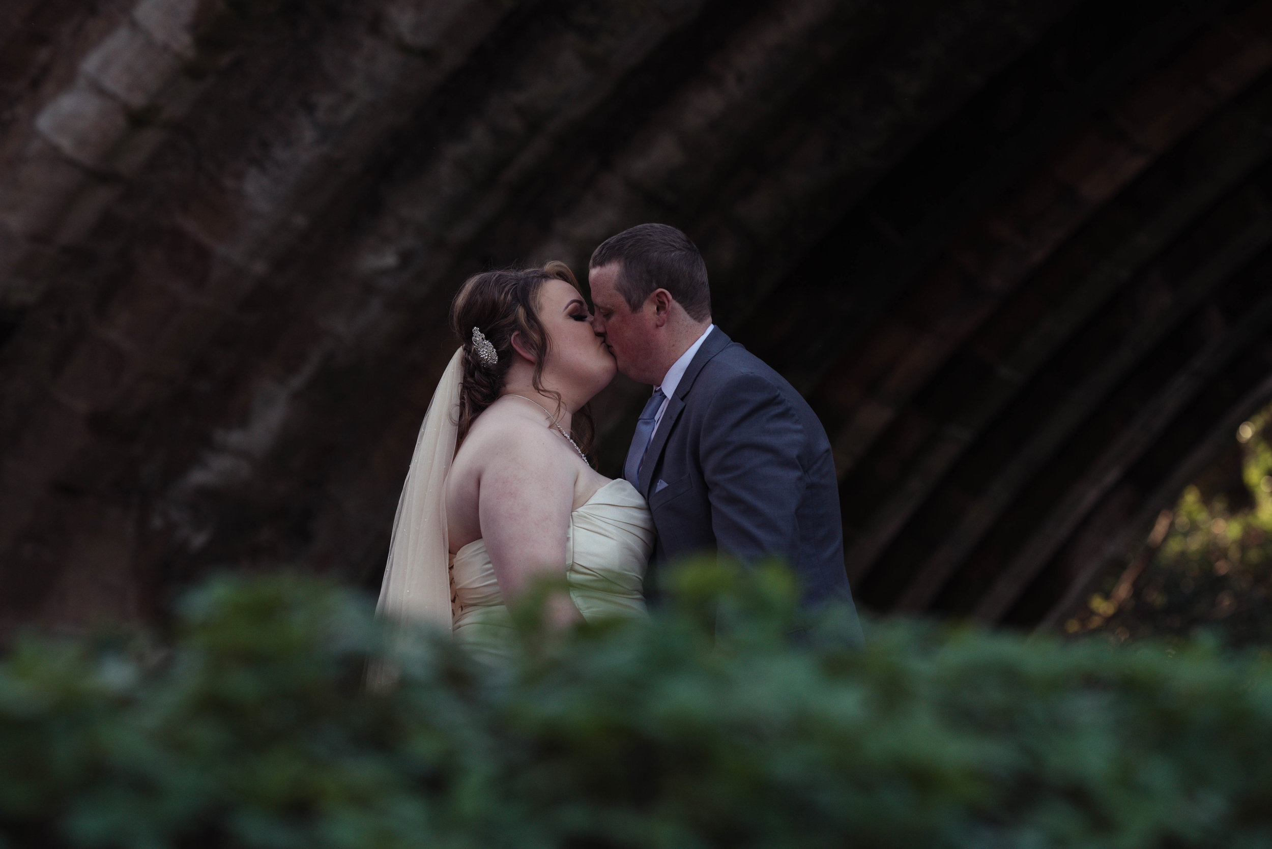 Partly hidden by a bush, the bride and groom share a kiss under a bridge