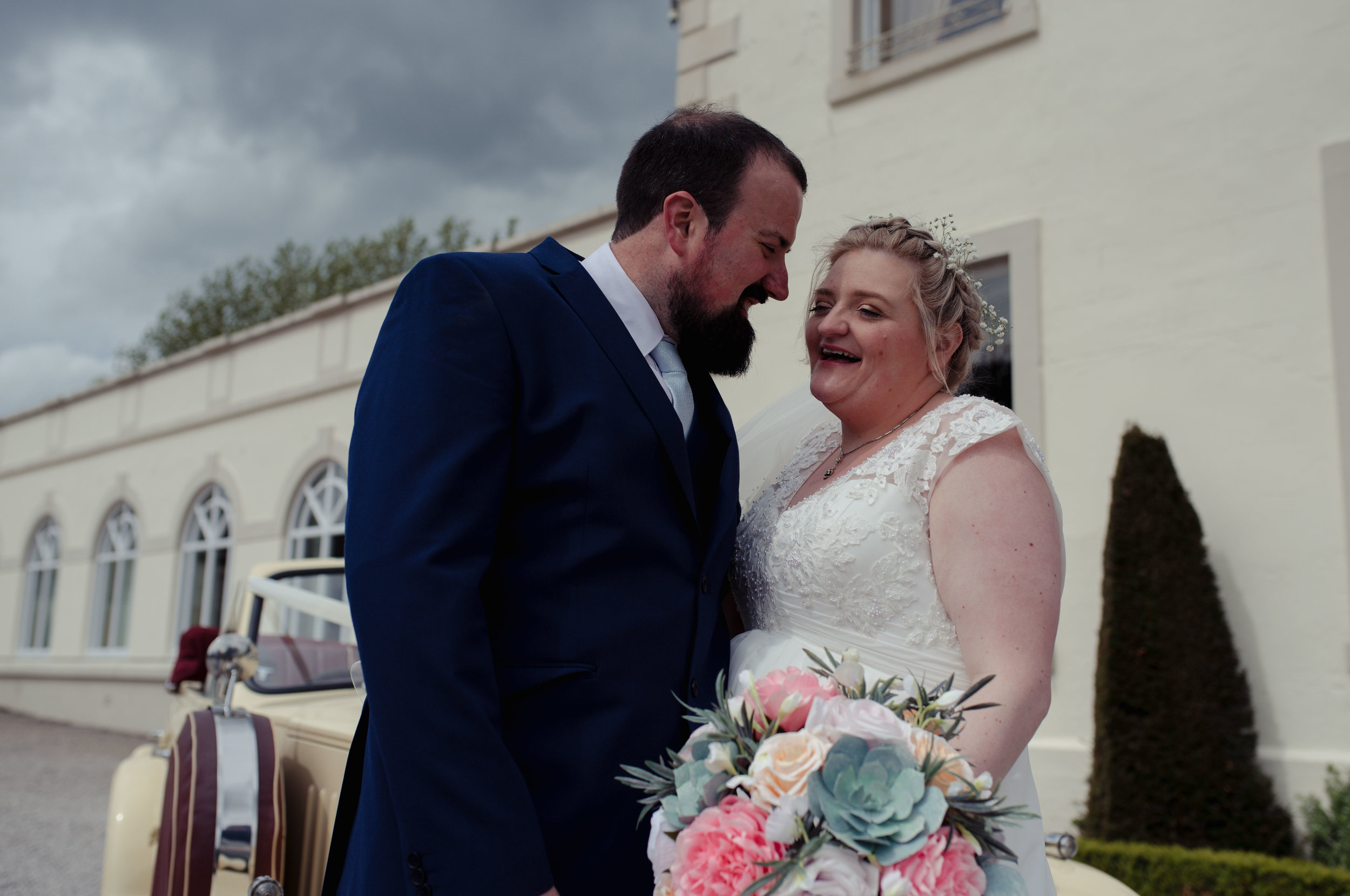 The bride and groom stand smiling next to their wedding car