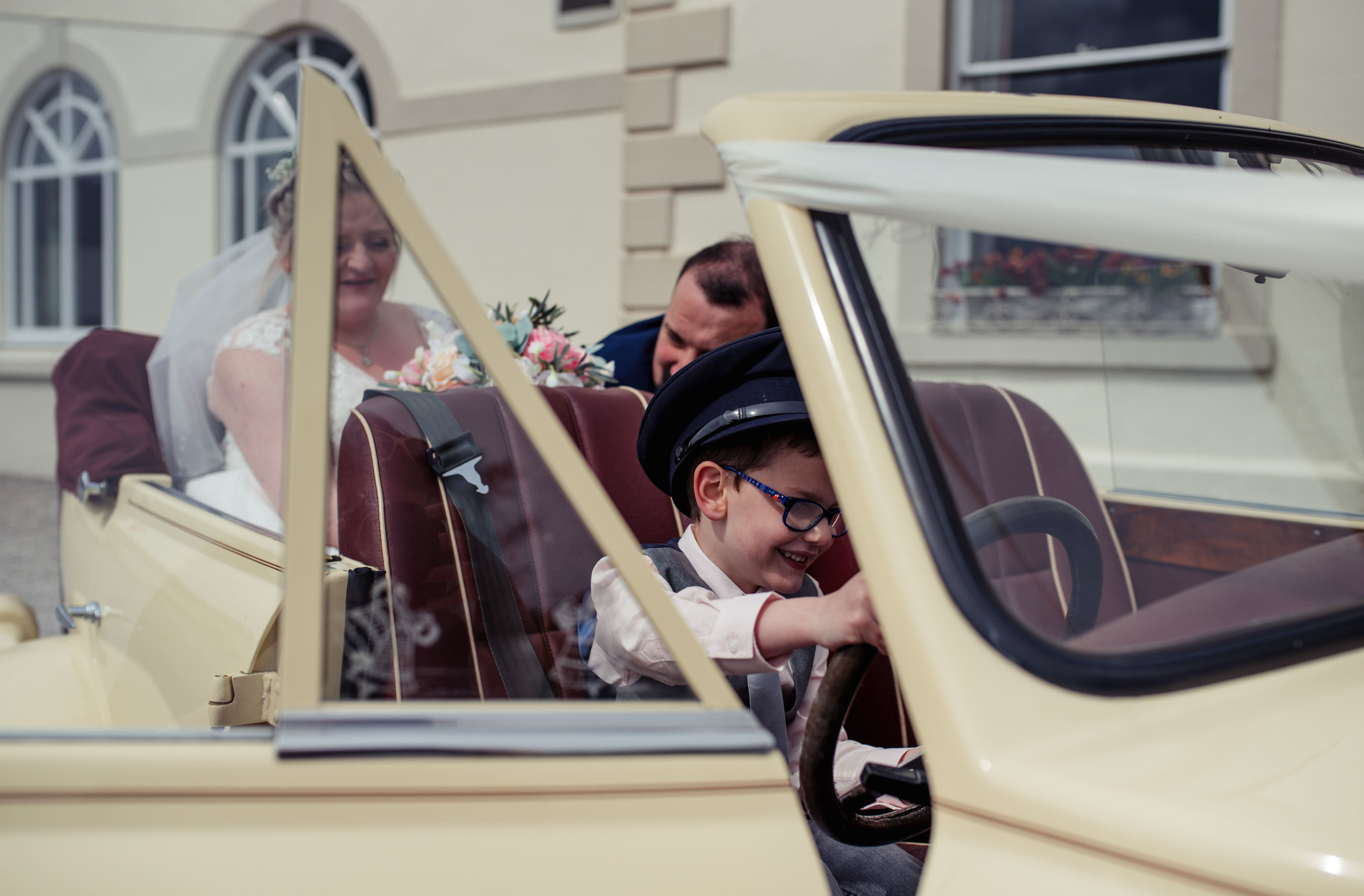 A page boys sits with a hat on in the drivers seat of the wedding car