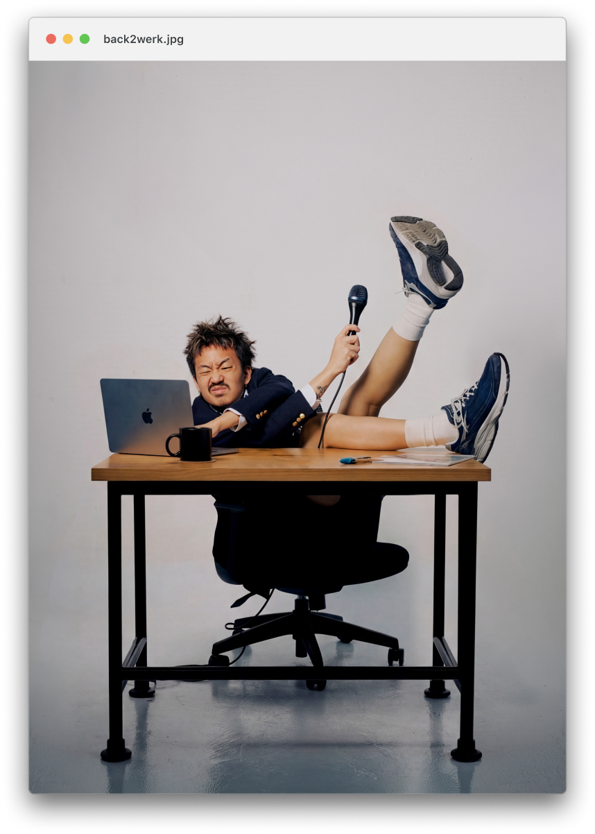 A person with short spiky hair lying on a desk with one leg raised, holding a microphone, with a laptop and a black mug on the desk, in front of a plain white background.