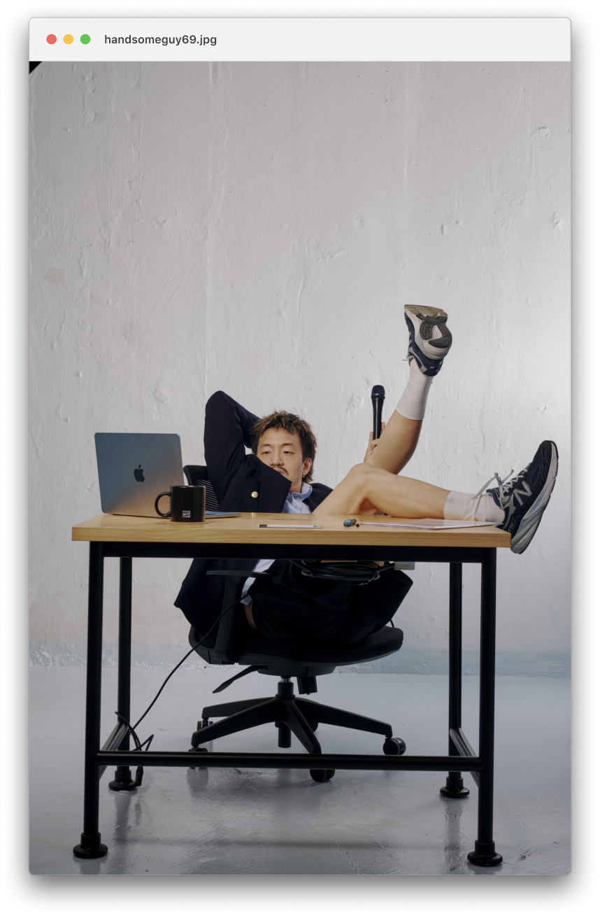 Jordan Leung dressed in business attire is sitting at a desk with one leg up, holding a microphone, with a laptop, mug, papers, and pen on the desk. The background is a plain white wall.