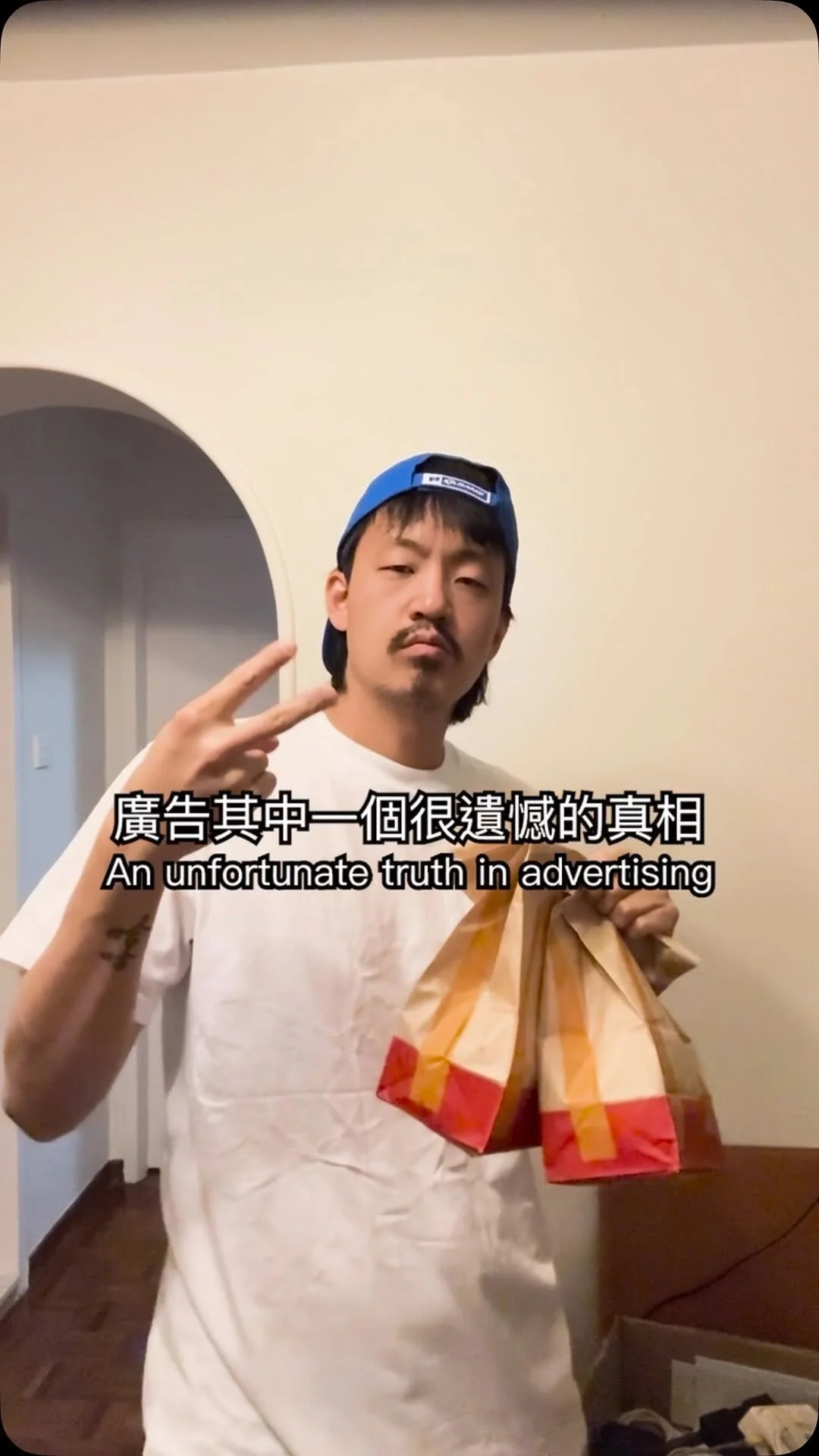 A man making a peace sign with his right hand, holding a paper bag in his left hand, standing in a room with light-colored walls and a door in the background, wearing a blue cap backwards and a white t-shirt.