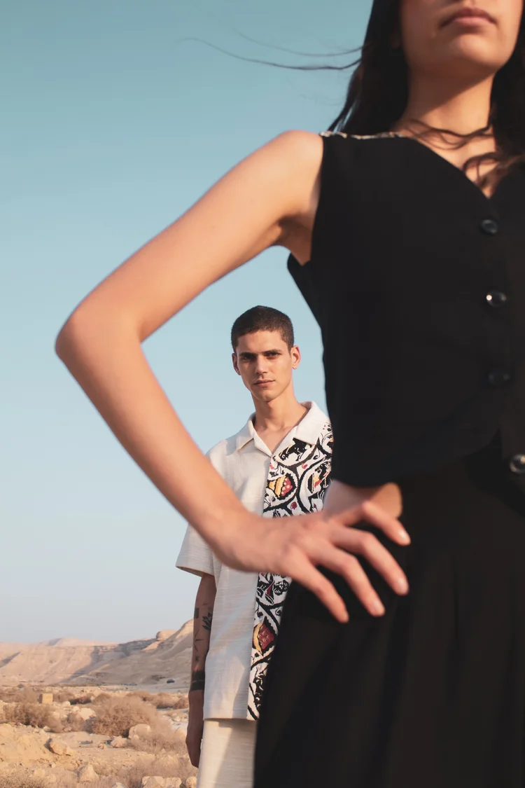 A woman in a black sleeveless dress with a man in a patterned shirt behind her, desert hills beyond