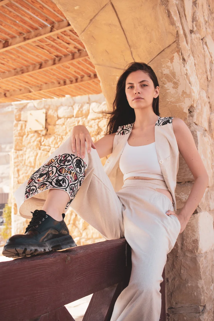 A woman in white with patterned trousers seated on a wooden bench under a pergola