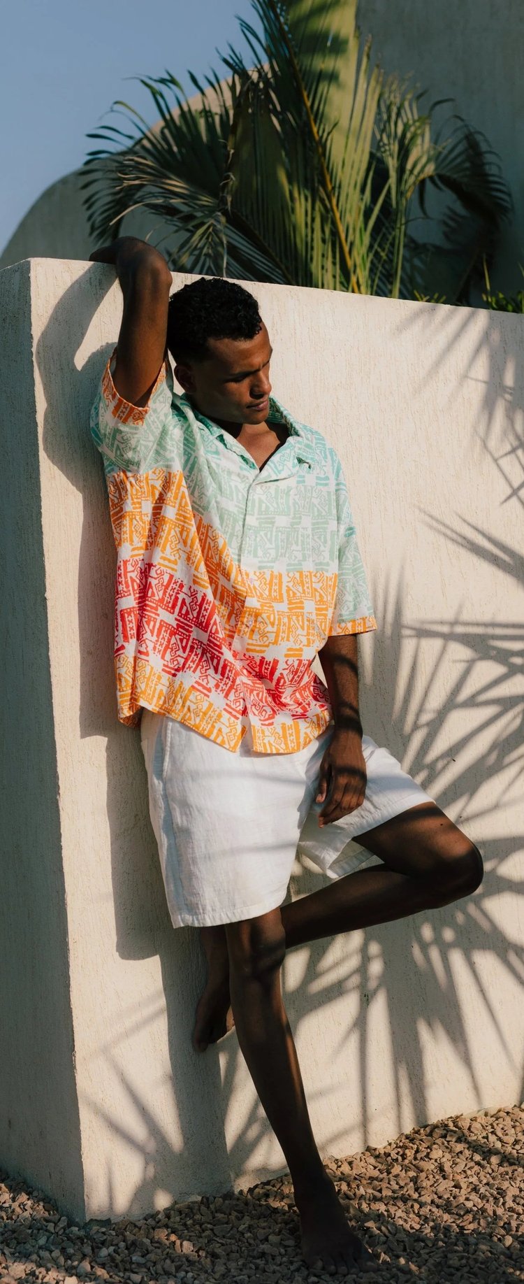 A man in a pink, orange and white patterned shirt leaning against a white wall