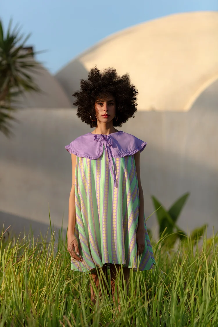 A woman in a striped dress with a lilac collar standing in tall grass before a white dome