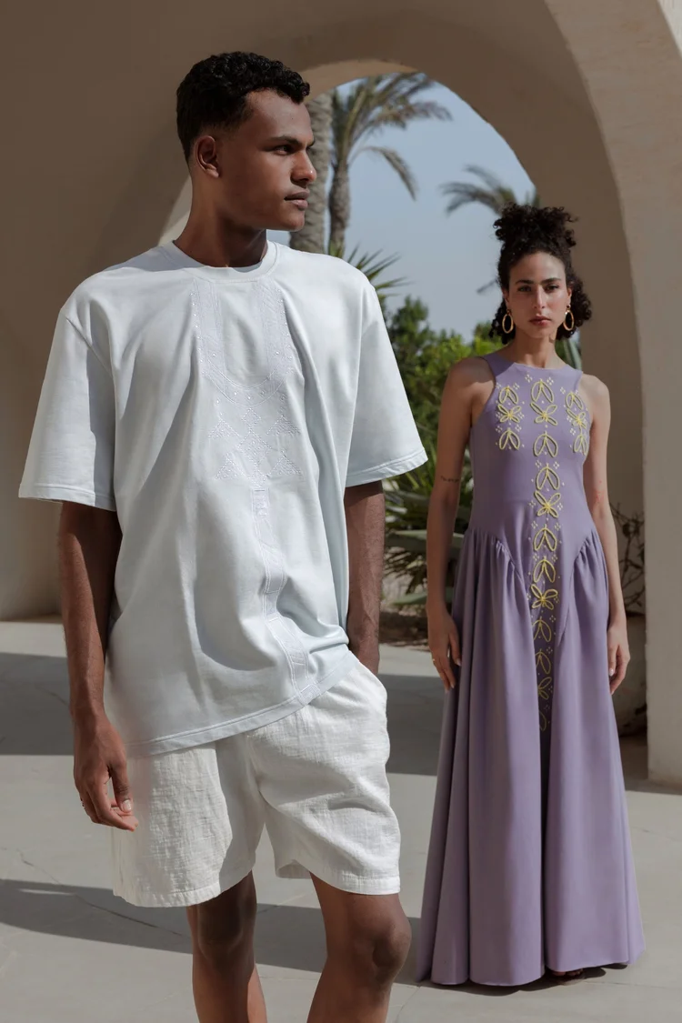 A man in a pale T-shirt and shorts and a woman in a lilac embroidered dress under a white arch with palms