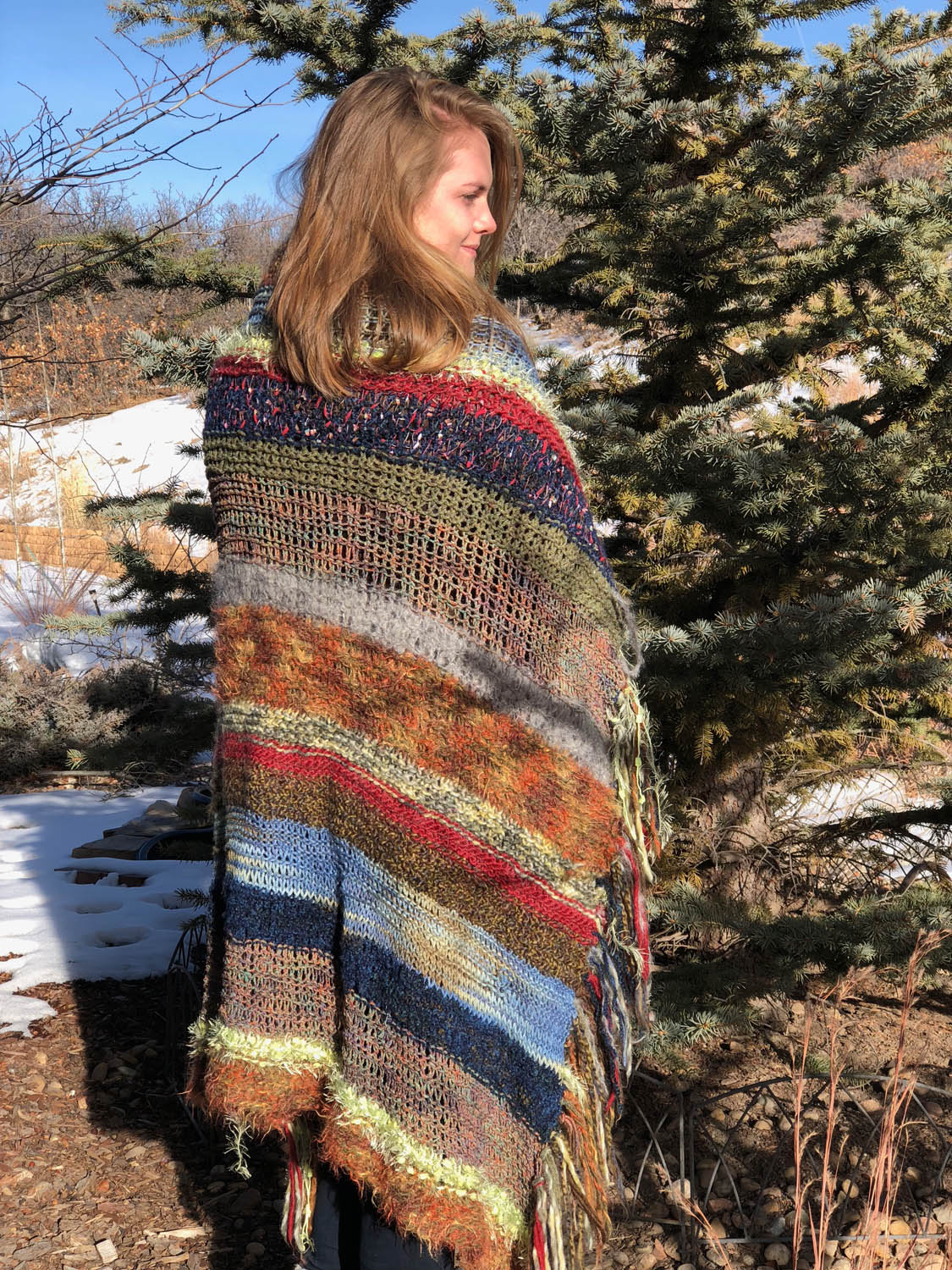 Model wearing scrap-yarn knitted throw as a shawl outside by pine tree in snow.