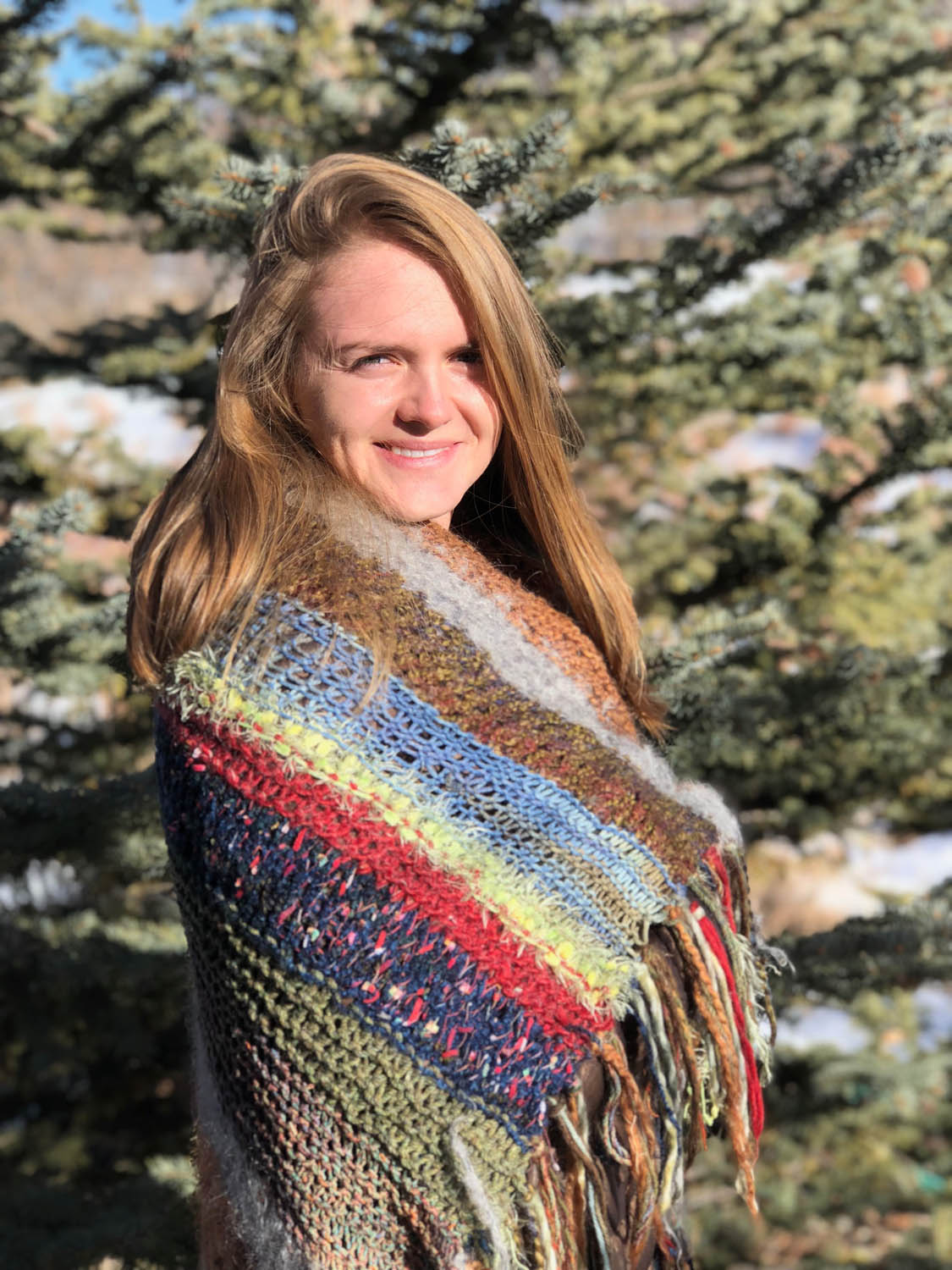 Young woman wrapped in scrap yarn knitted throw outdoors with pine tree and snow.