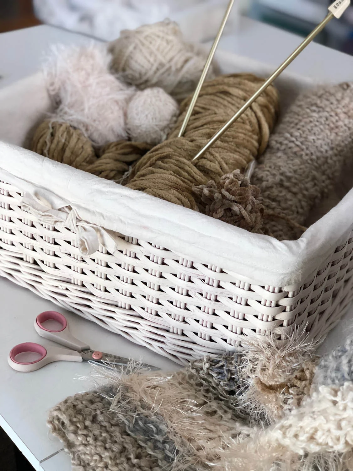 White wicker basket filled with variety of yarn balls in neutral winter whites with knitting needles