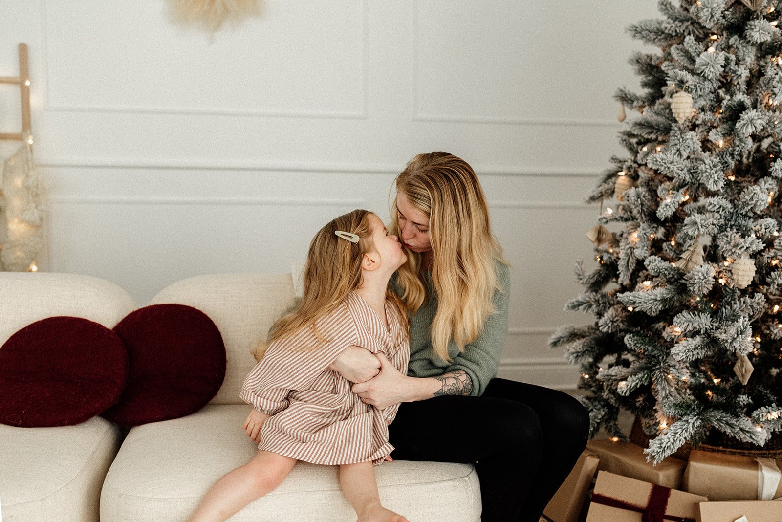 little girl kissing her mom sitting beside Christmas tree in a Toronto studio