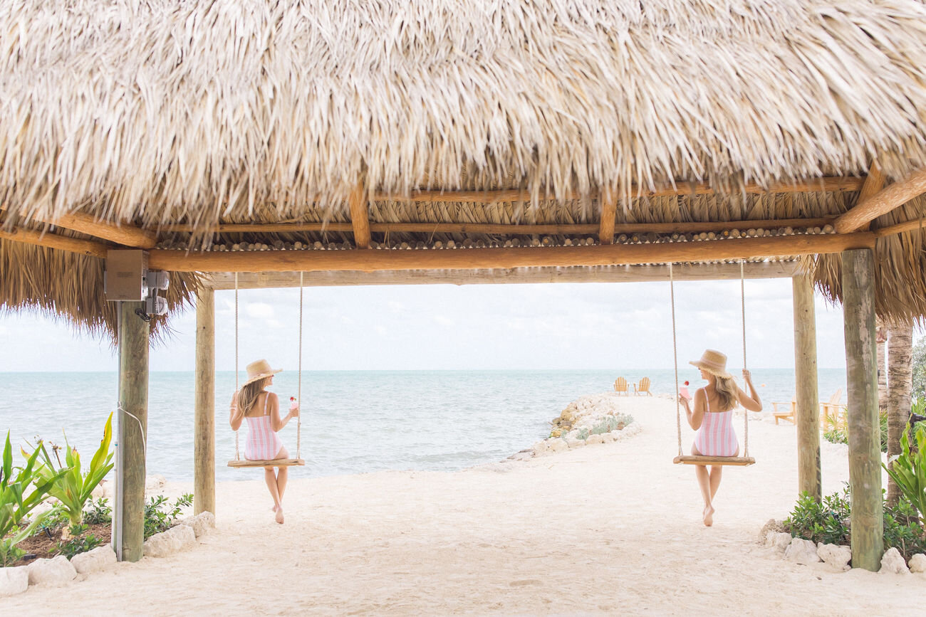  Two women in matching swimsuits sitting on swings under The Islands of Islamorada’s private beach hut. 
