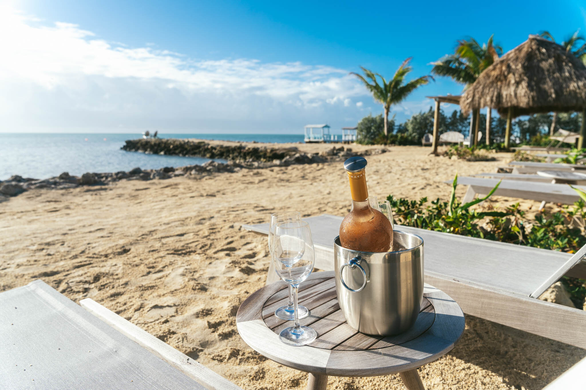  Bottle Of Wine In An Ice Bucket With Two Glasses On A Table Next To Two Lounge Chairs On The Islands Of Islamorada’s Private Beach. 