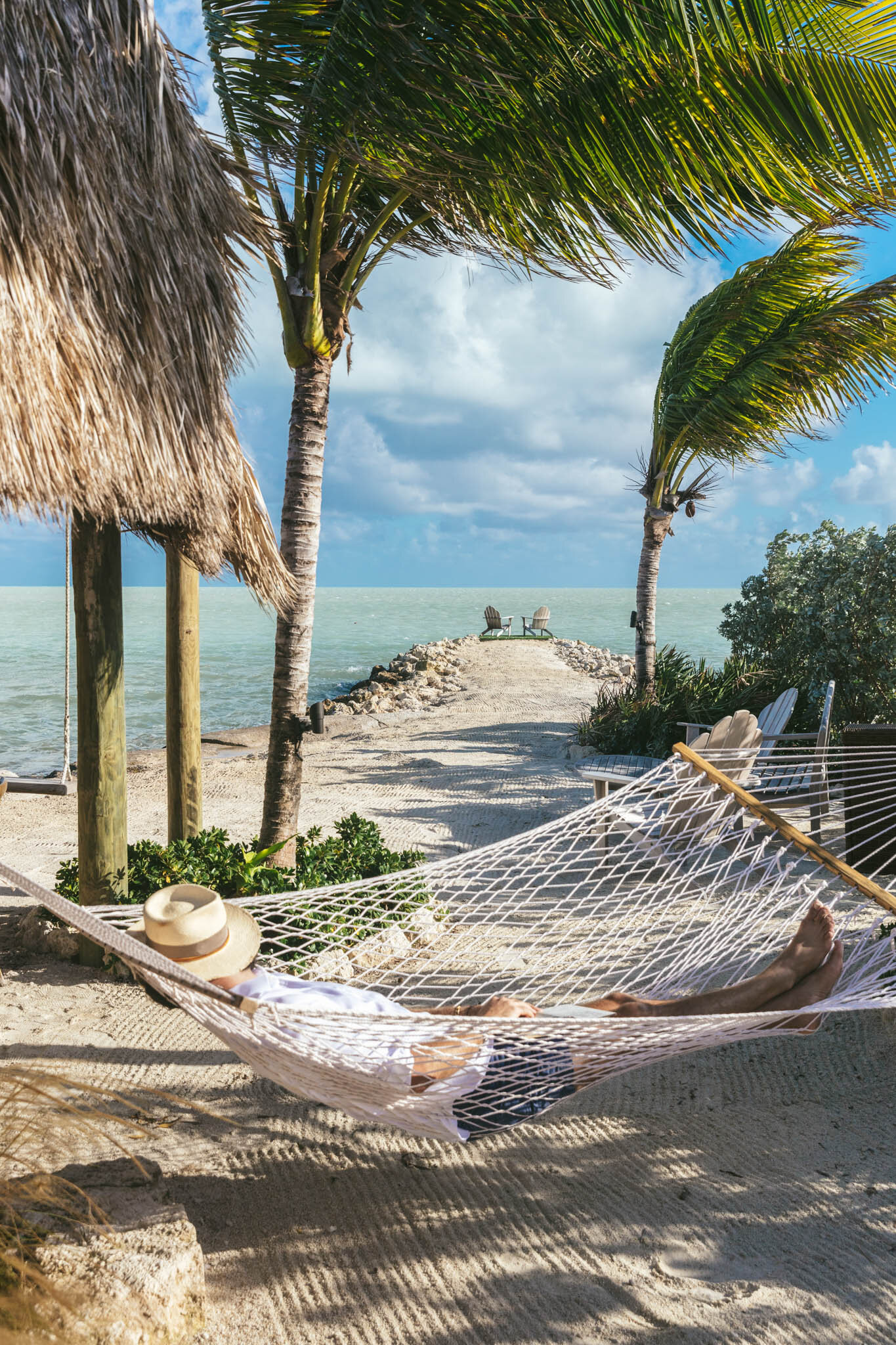  Man lying on a hammock with a hat over his face next to The Islands of Islamorada’s tiki bar hut, with the resort’s private jetty looking out onto the Atlantic Ocean in the background. 
