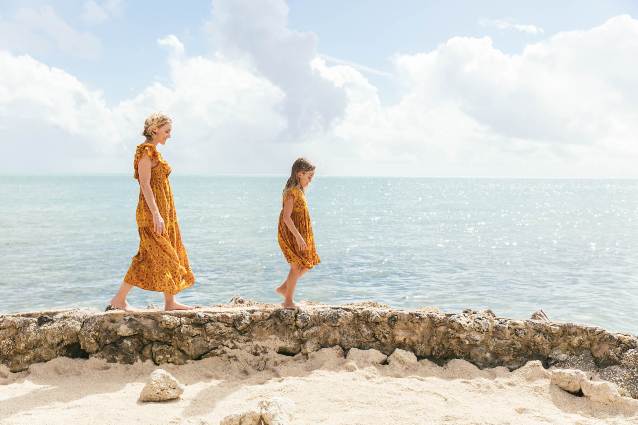  Mother and young daughter in matching summer dresses walk along the rocky ledge on The Islands of Islamorada’s private beach. 