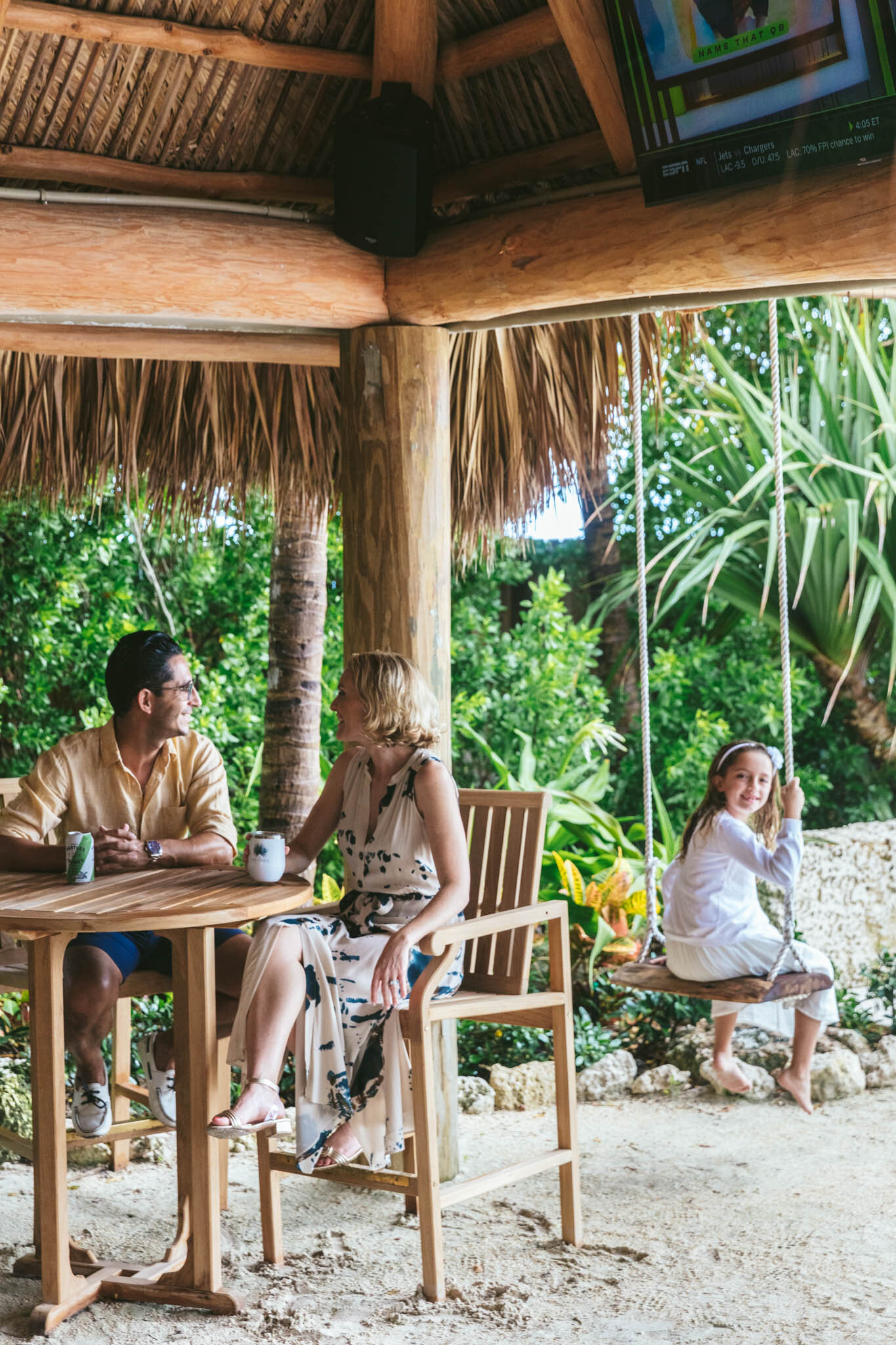  Young girl sitting on a swing under The Islands of Islamorada’s beachside tiki bar hut while her mom and dad sit and watch her from a nearby table. 