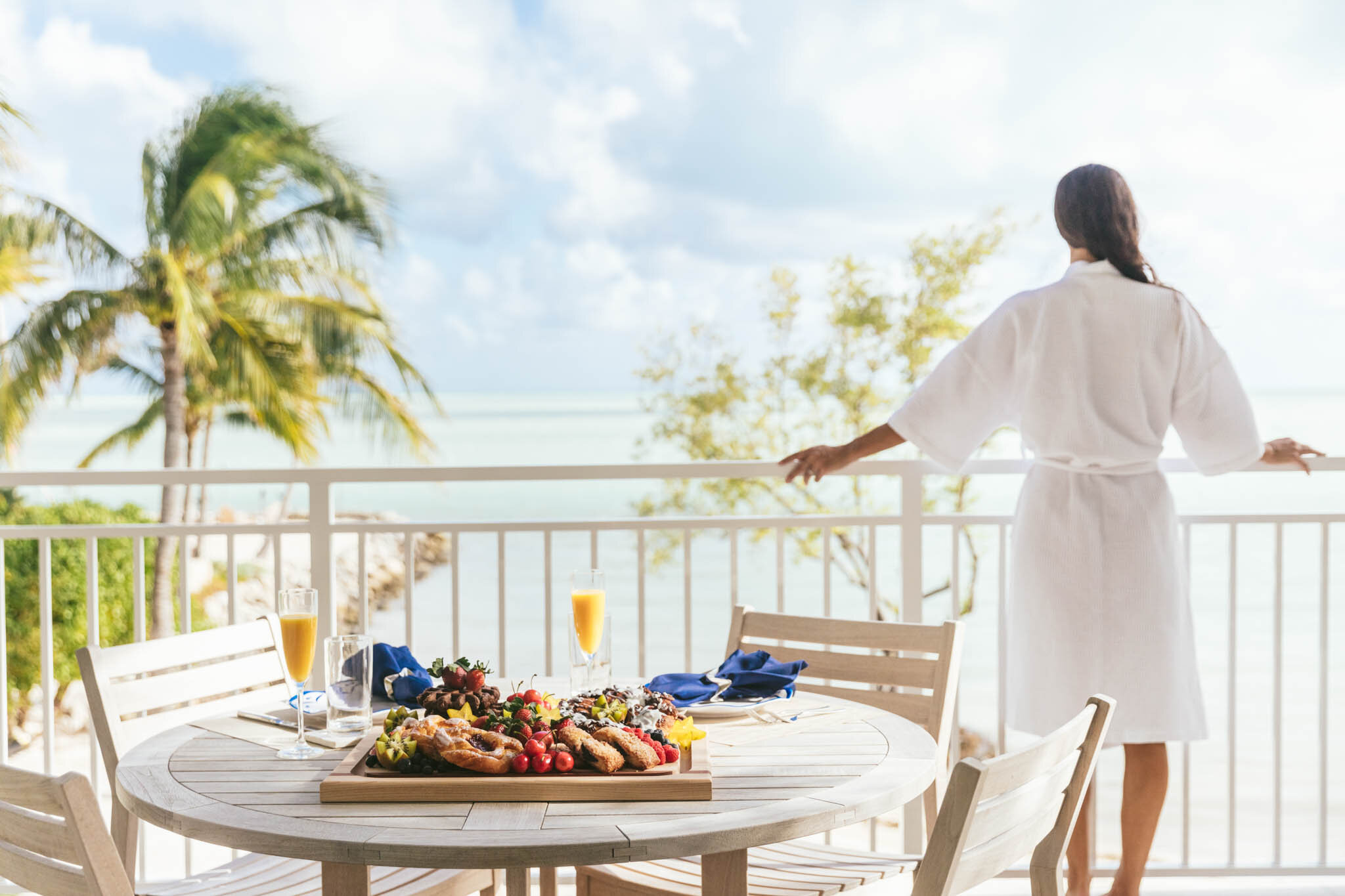  Woman in a bath robe stands on the balcony of an Islands waterfront villa looking out onto the Atlantic Ocean behind a table set for breakfast. 