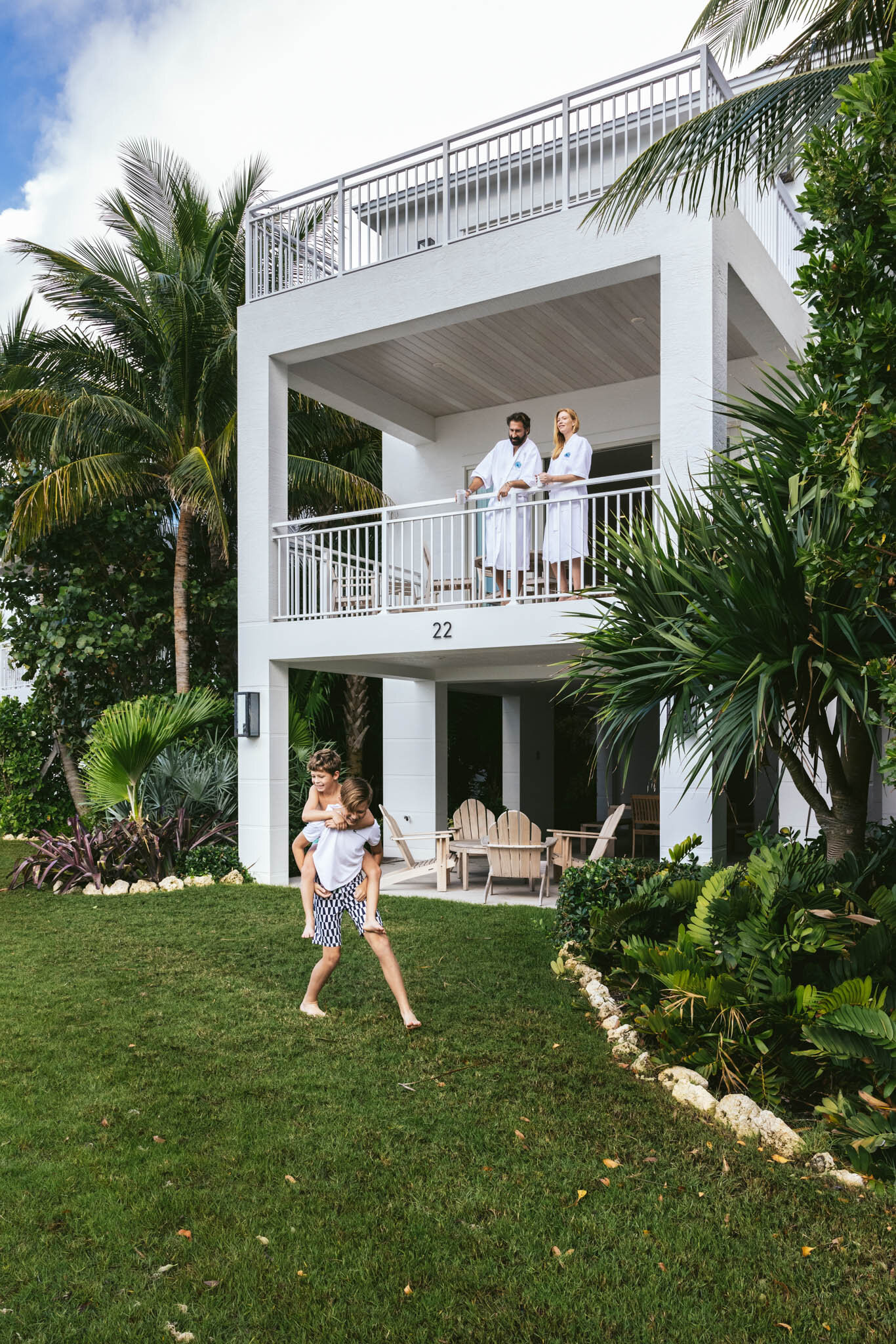 Two kids playing on the open lawn in front of an Islands waterfront villa as their parents look on from a covered first floor balcony. 