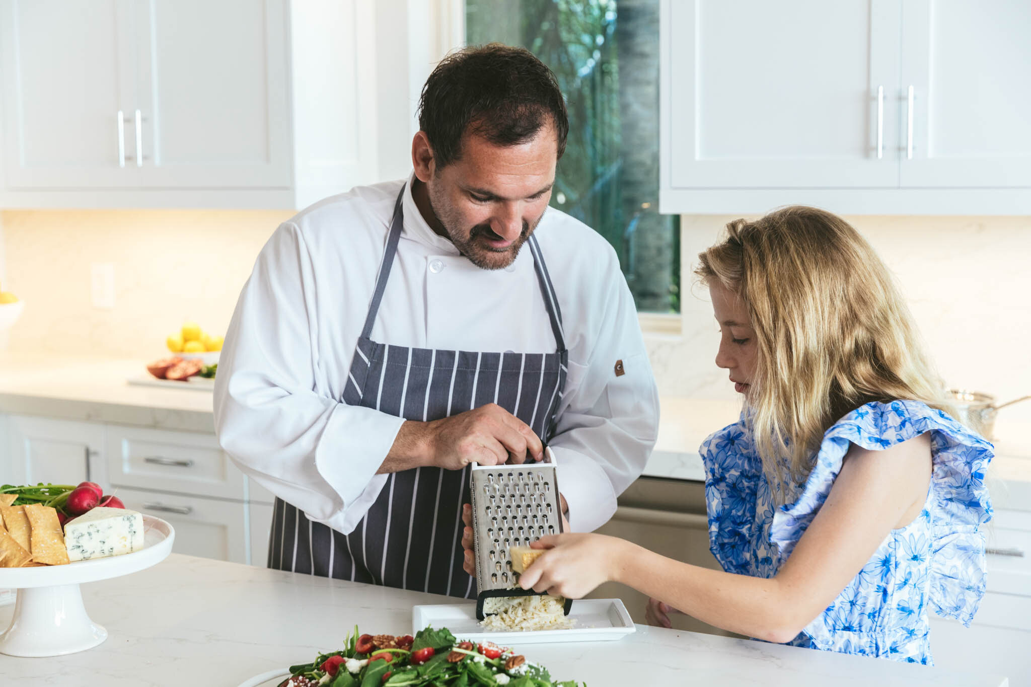  A chef lets a young girl grate cheese while preparing a meal in the kitchen of an Islands waterfront villa. 