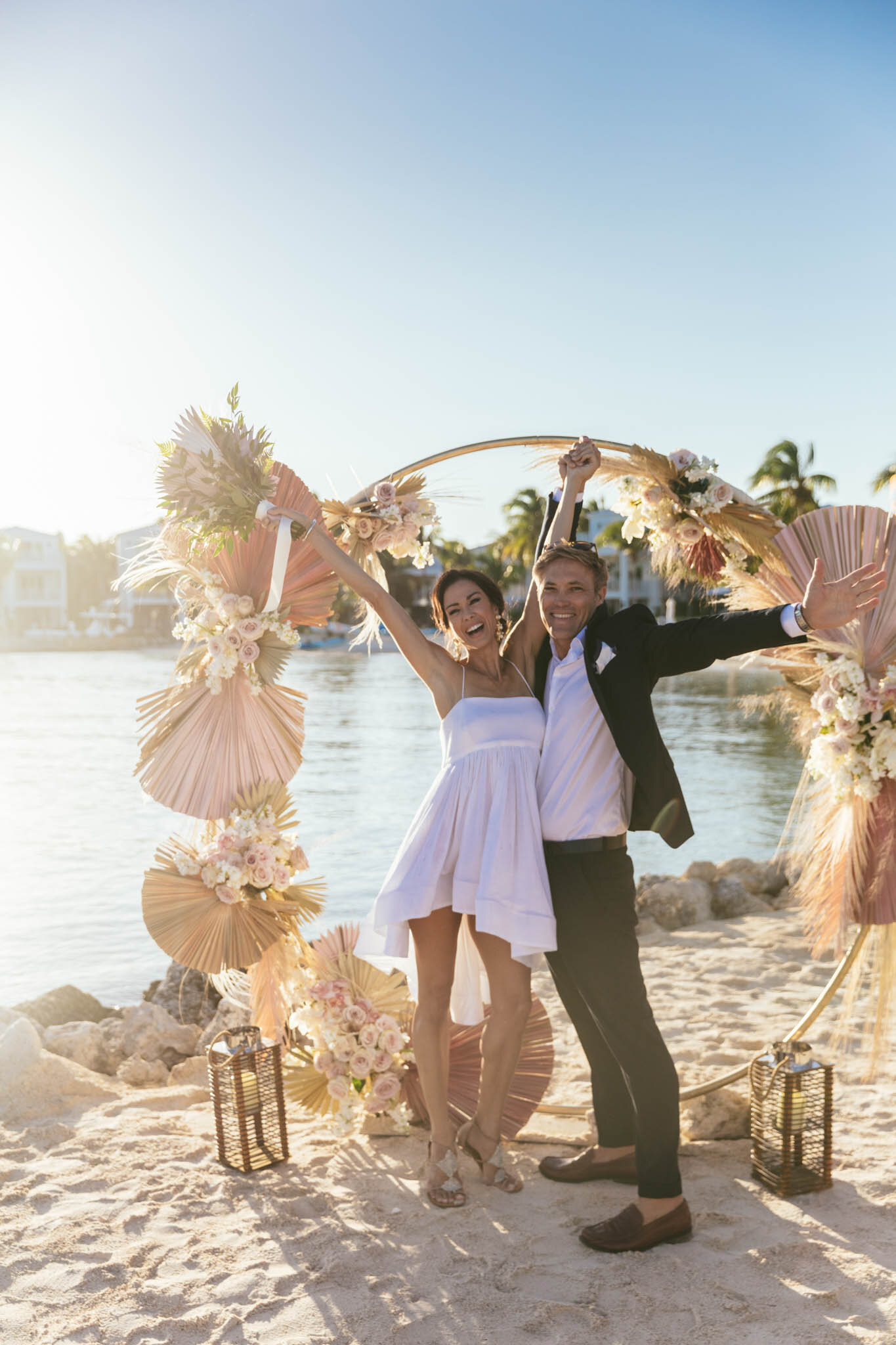  Bride and groom cheer with their arms in the air under a wedding arch on The Islands of Islamorada’s private beach. 