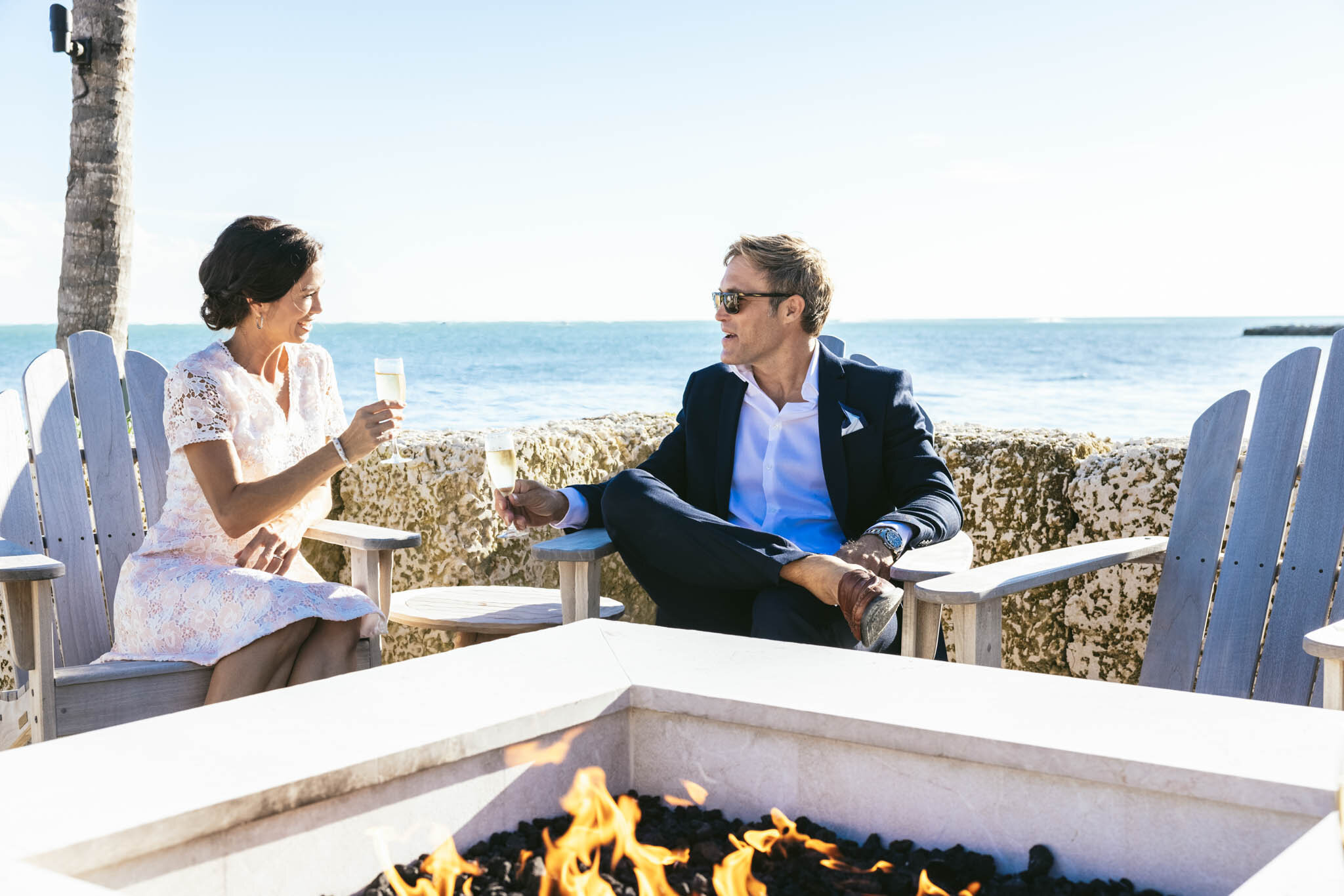  Bride and groom raise glasses of champagne together while sitting by The Islands of Islamorada’s fire pit on the beach. 