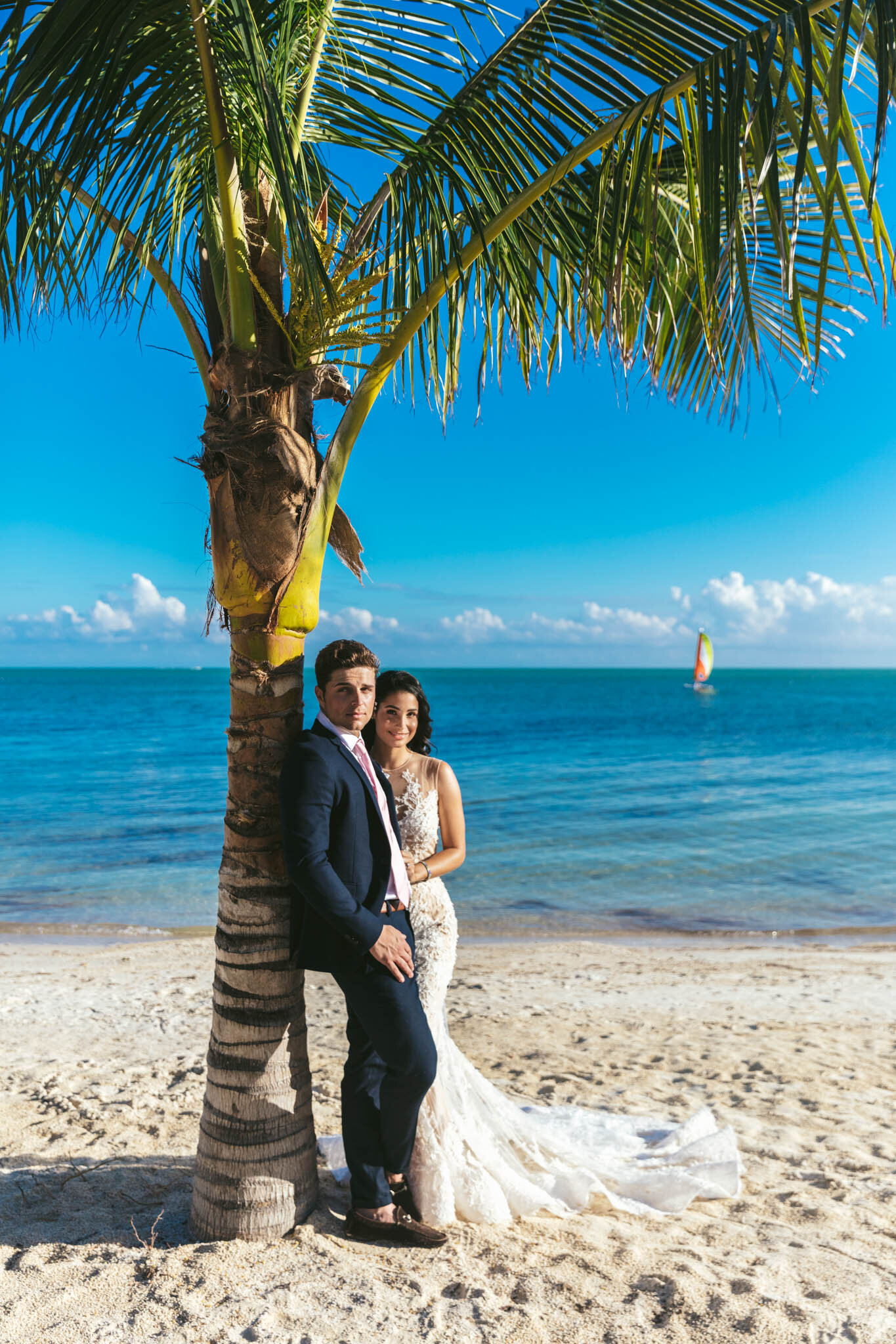  Bride and groom stand together under a palm tree on The Islands of Islamorada’s private beach with a backdrop of the Atlantic Ocean and a sailboat in the distance. 
