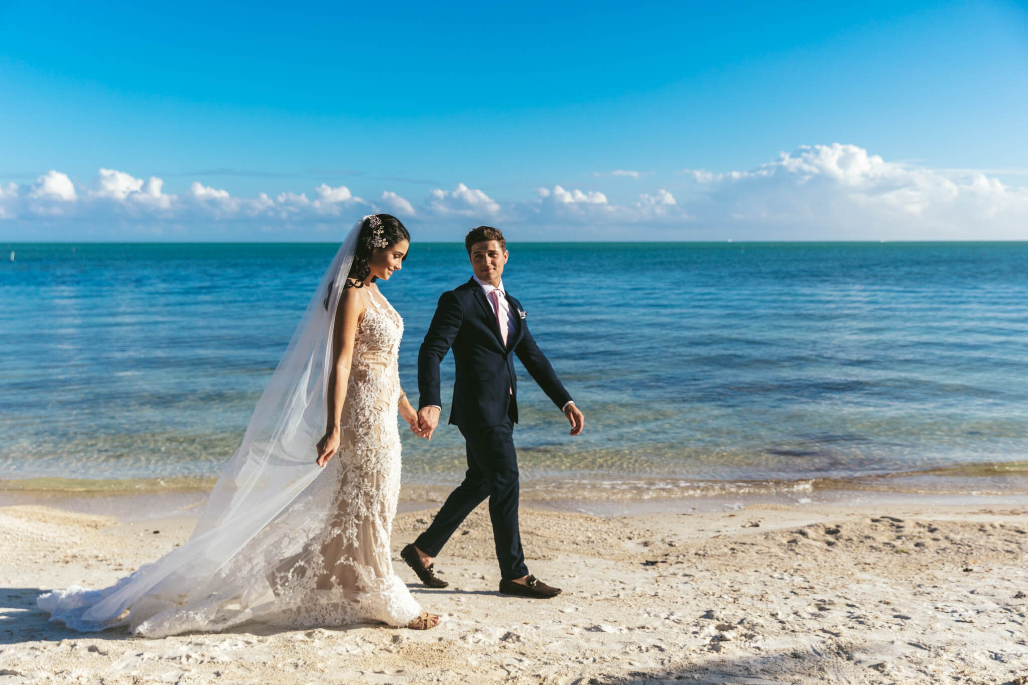  Bride and groom walking together and holding hands on The Islands of Islamorada’s private beach with the Atlantic Ocean as a backdrop. 