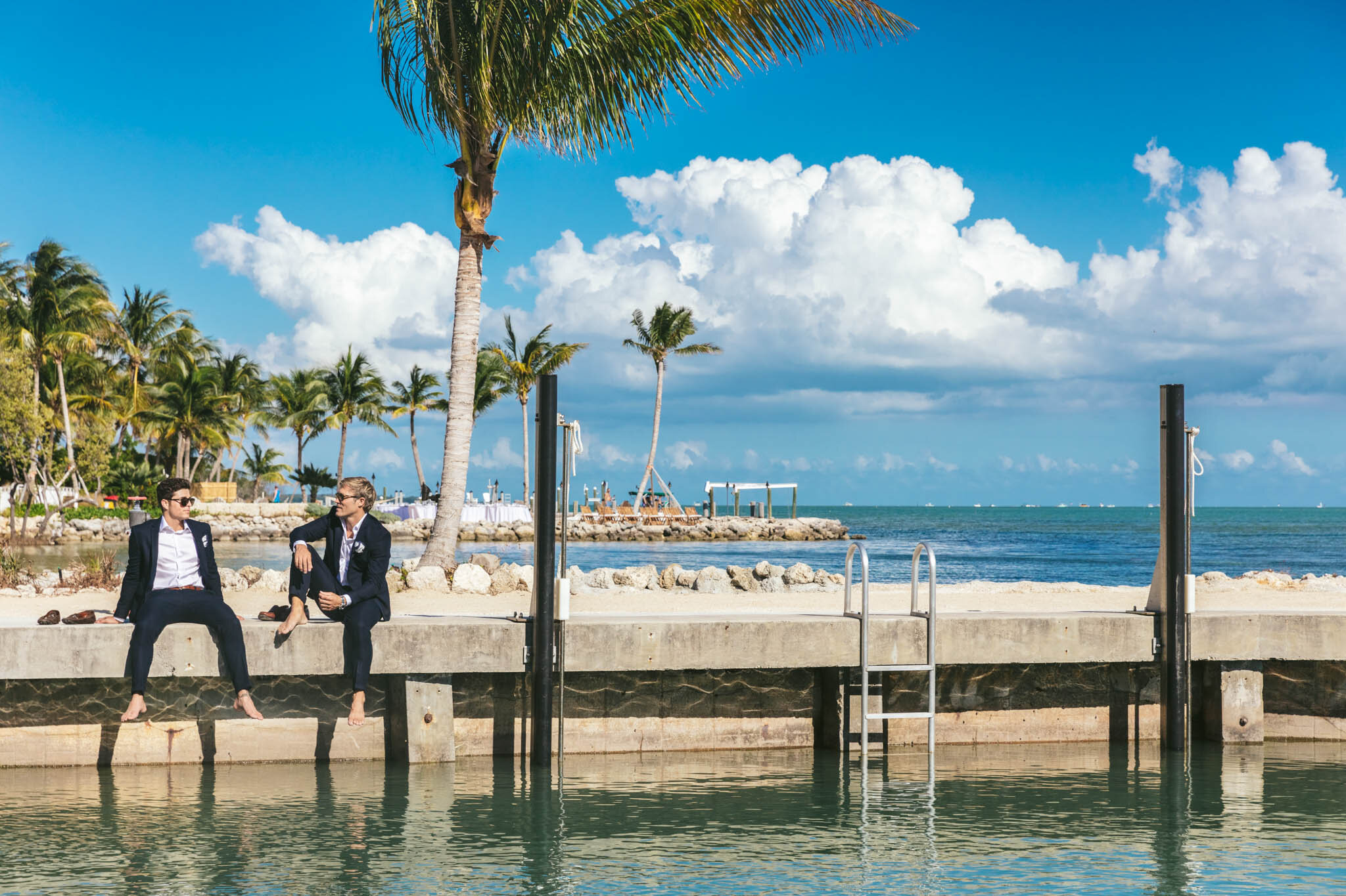  Groom and best man in wedding tuxedos sitting with their shoes off on the side of The Islands of Islamorada’s private marina dock. 