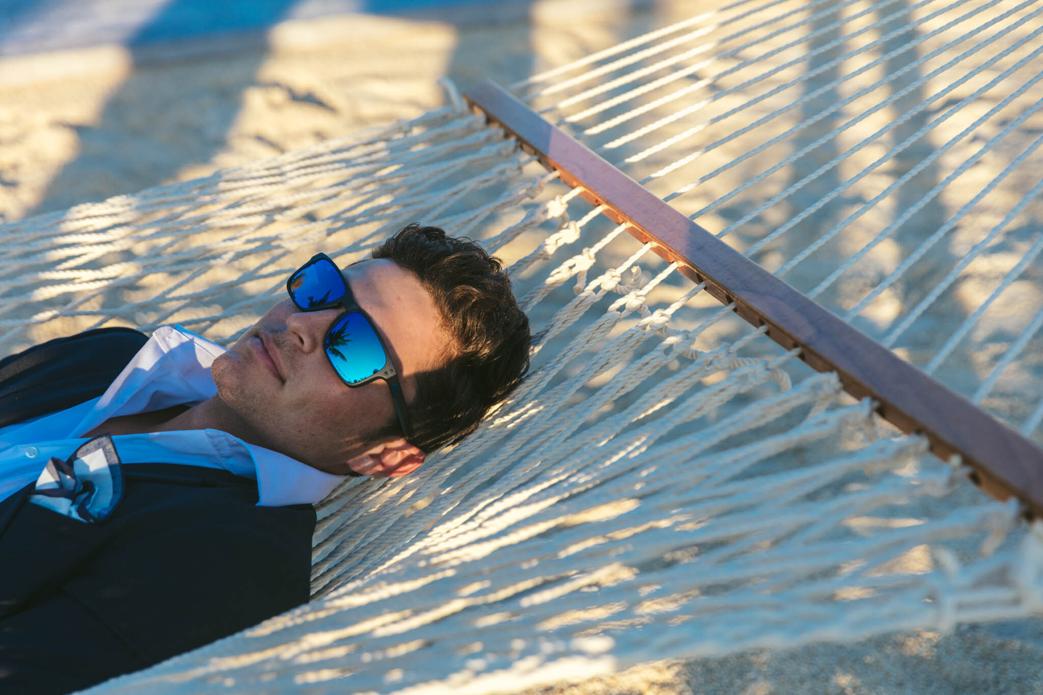  Groom wearing sunglasses and a wedding tuxedo lies relaxing on a hammock on The Islands of Islamorada’s private beach. 