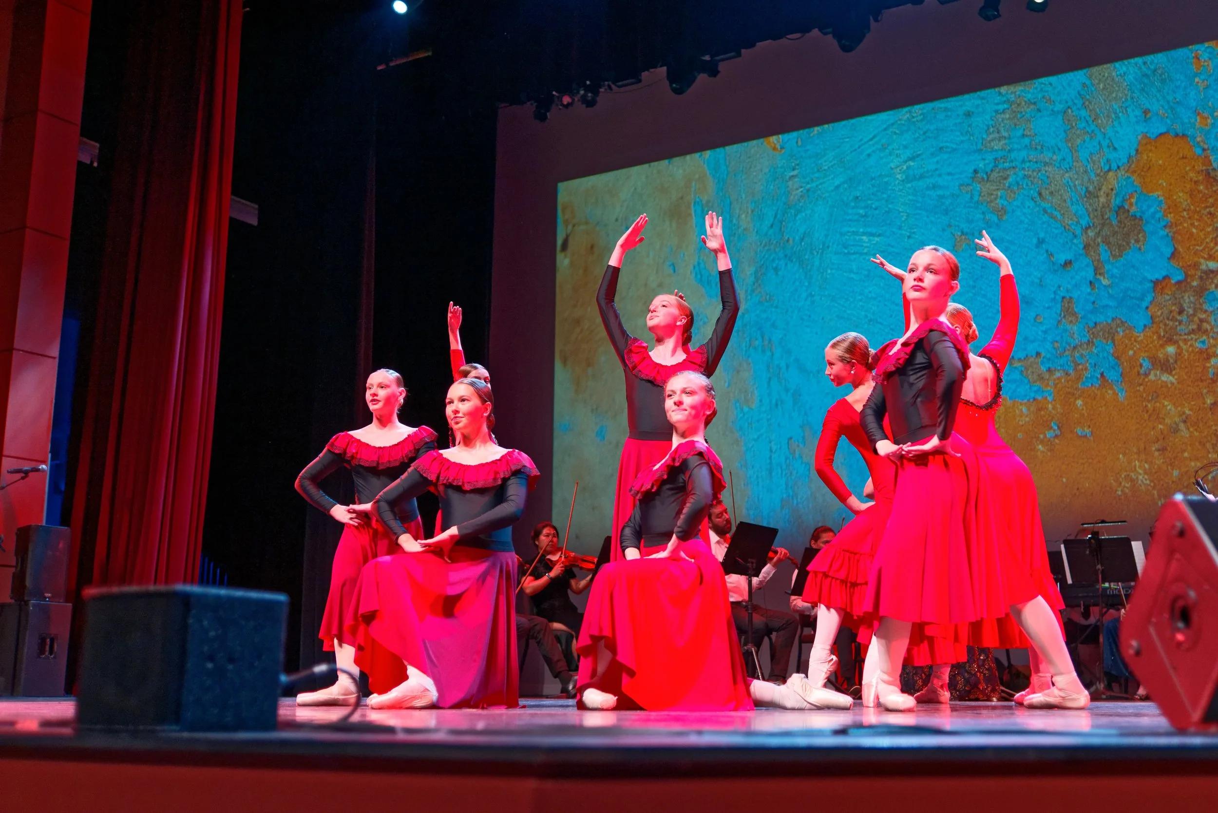 A group of young ballet dancers in red and black costumes performing on stage with musicians in the background and a colorful backdrop.