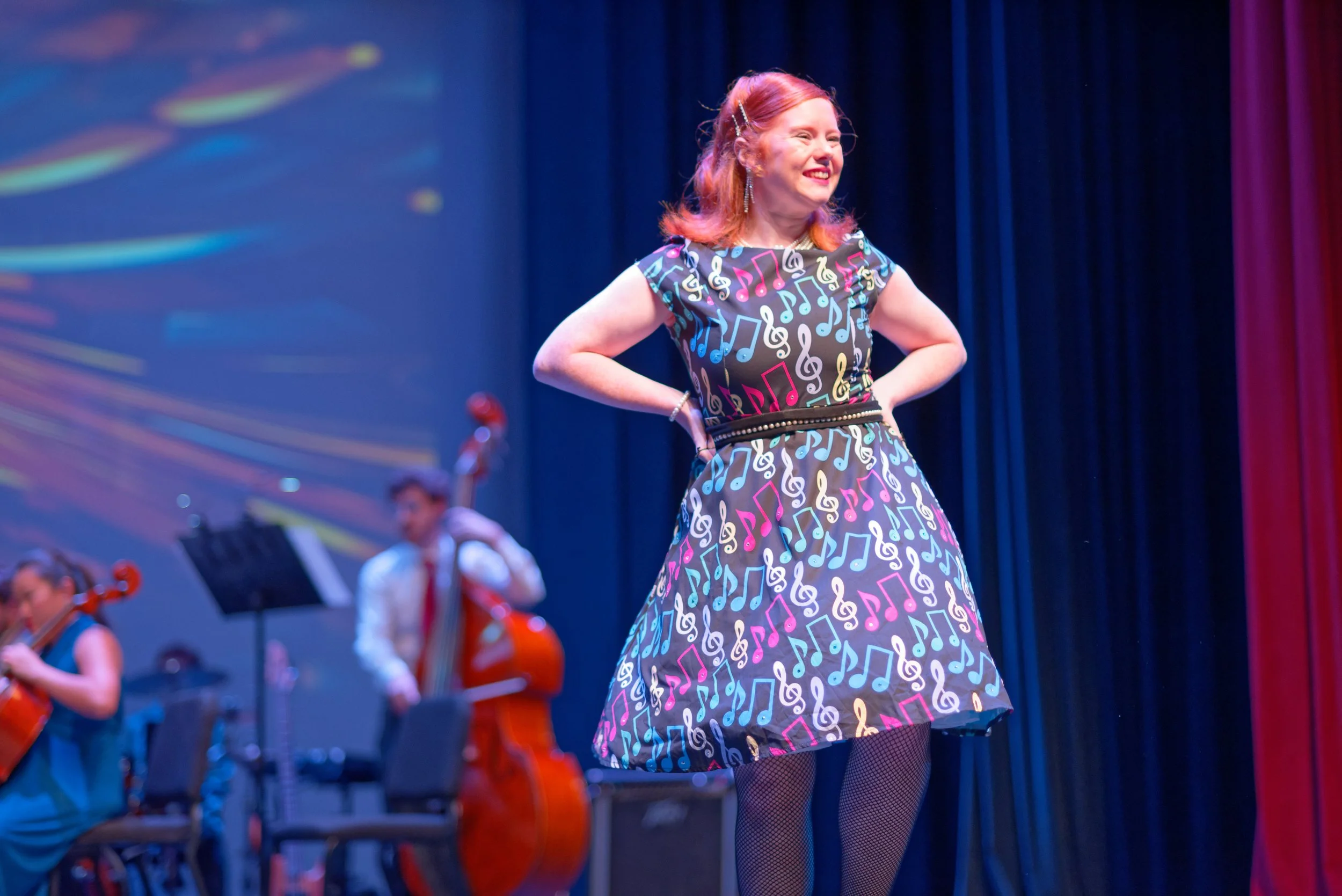 A woman with red hair, dressed in a colorful dress with musical notes, performing on stage with an orchestra in the background.