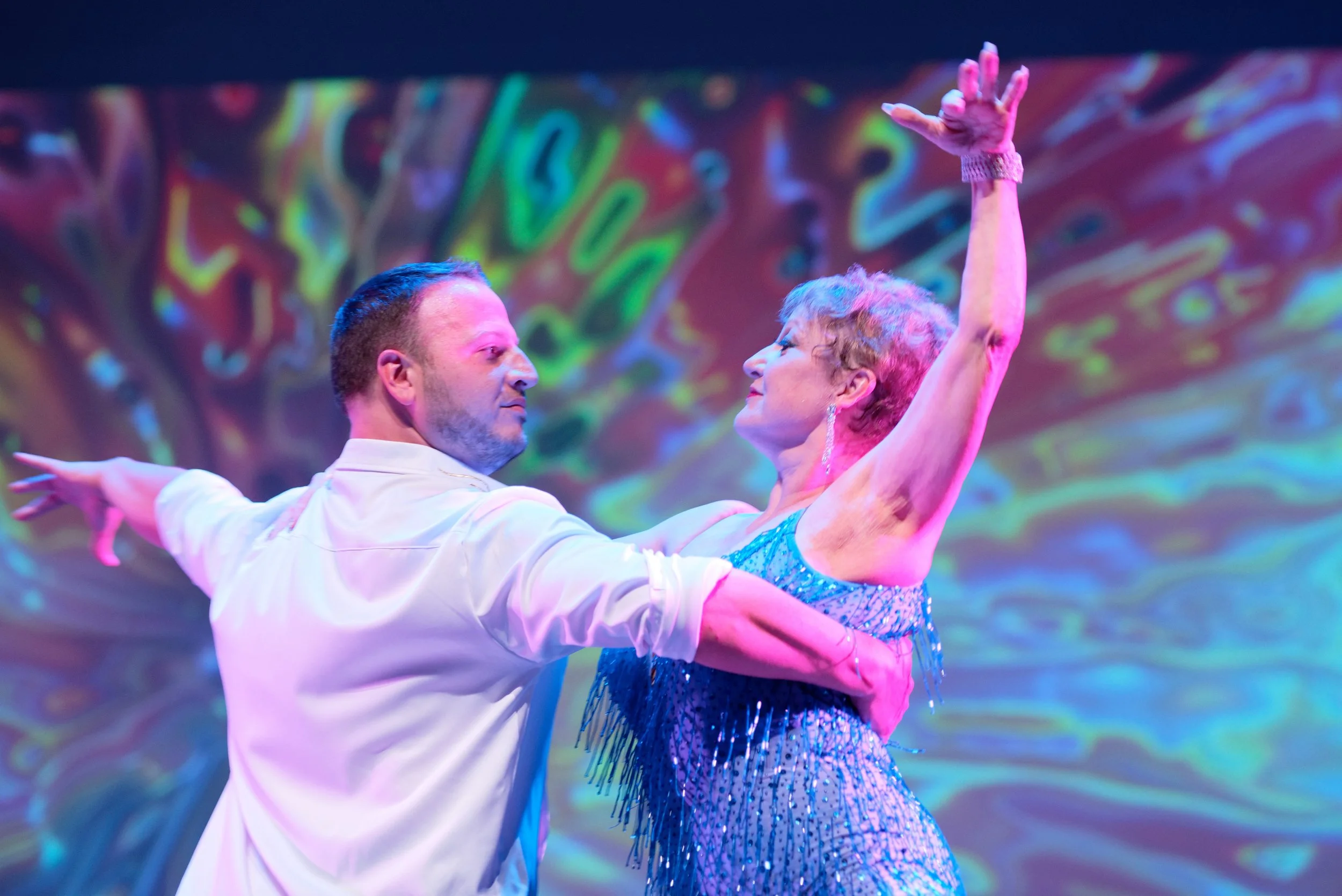 A man and woman dancing ballroom style with a colorful, abstract background. The woman is wearing a blue dress with fringe and the man is in a white shirt.