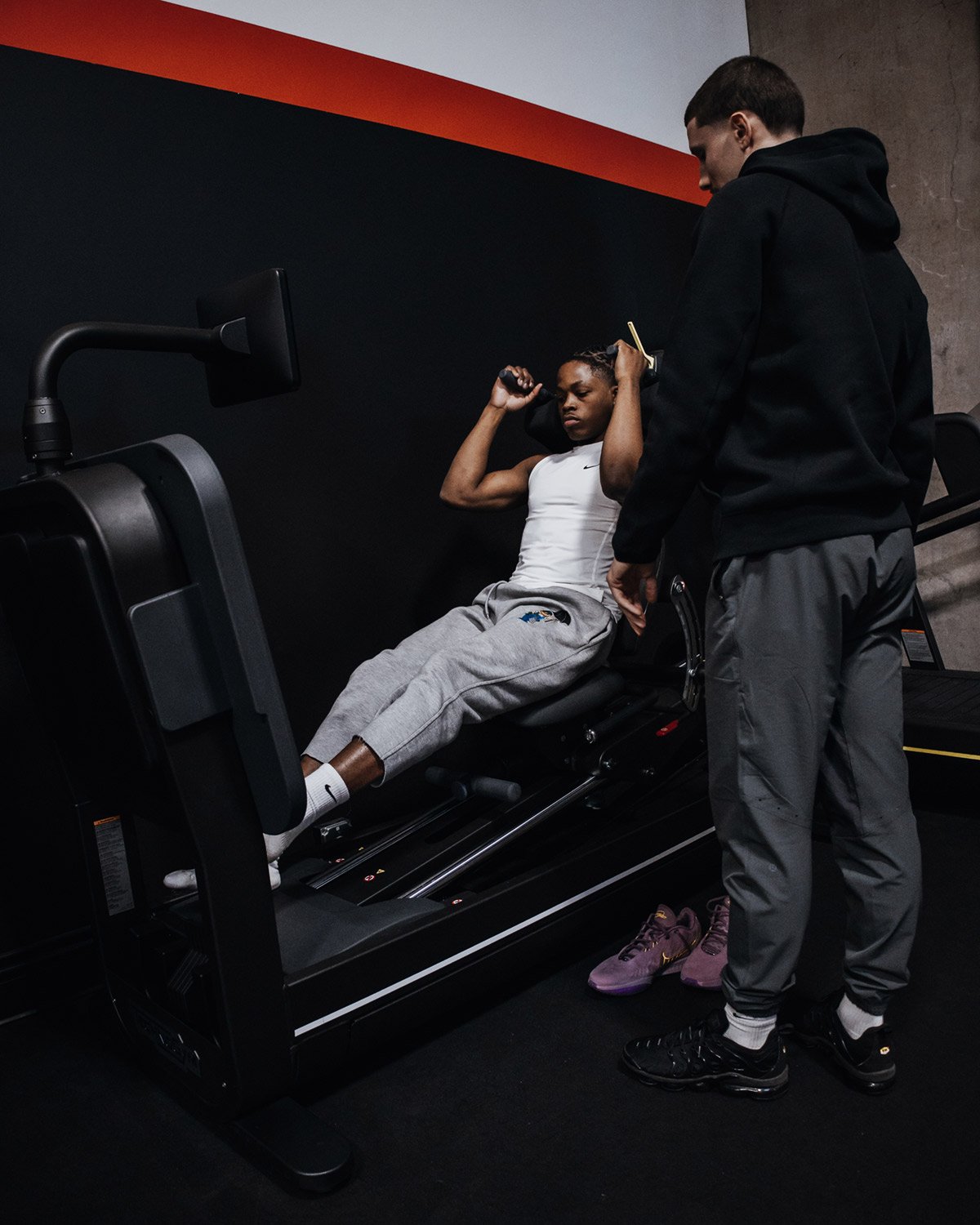 Two people in a gym with one using an exercise machine and the other standing next to them for assistance.