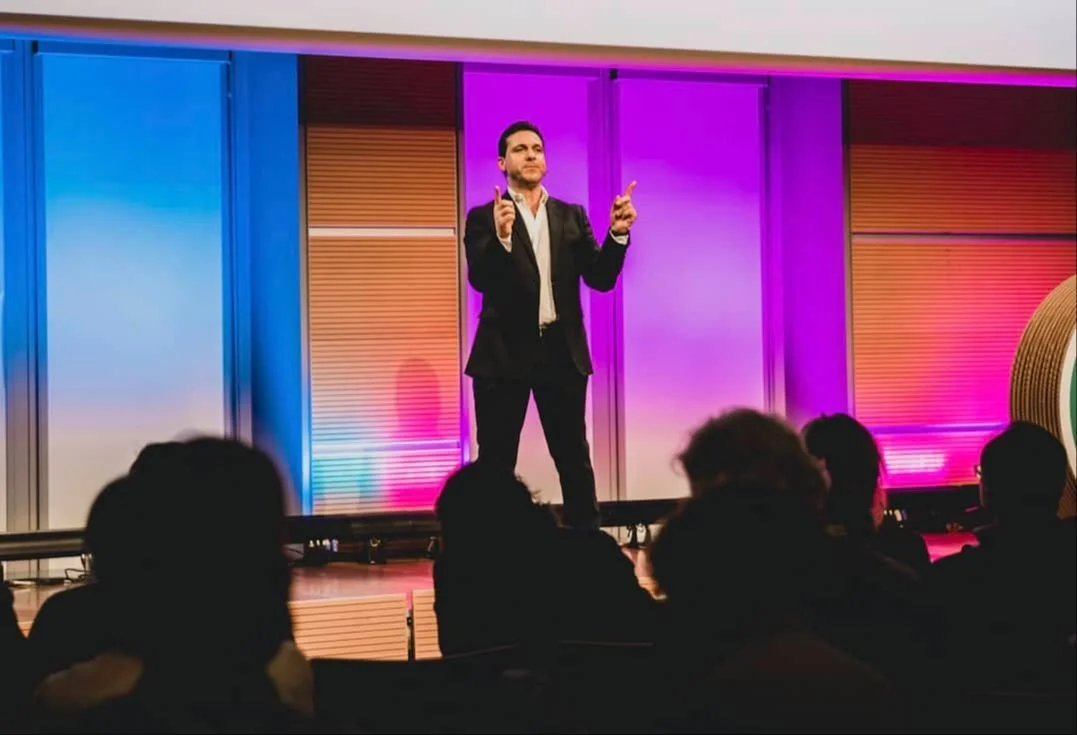 Uomo in un completo nero che parla davanti a un pubblico seduto, sul palco con sfondo colorato rosa e blu