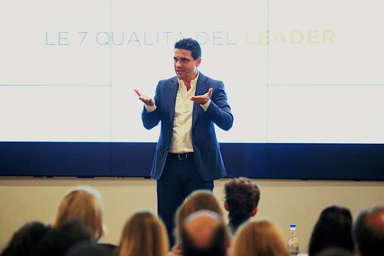 Un uomo in abito blu che parla davanti a un pubblico in una presentazione, con uno schermo alle spalle che dice "Le 7 qualità del leader".
