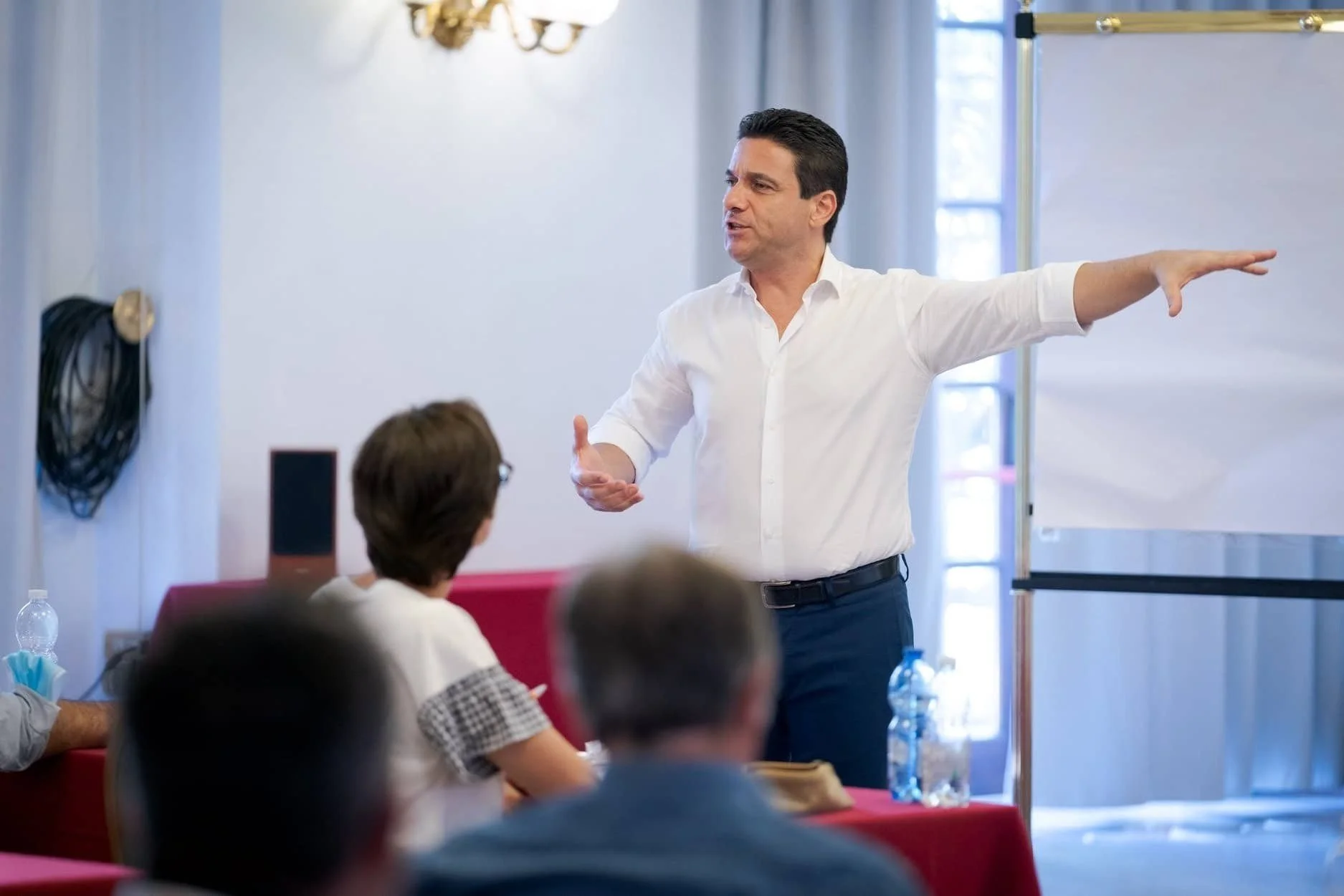 Uomo in camicia bianca che parla e fa gesture durante una presentazione davanti ad un pubblico in un'aula