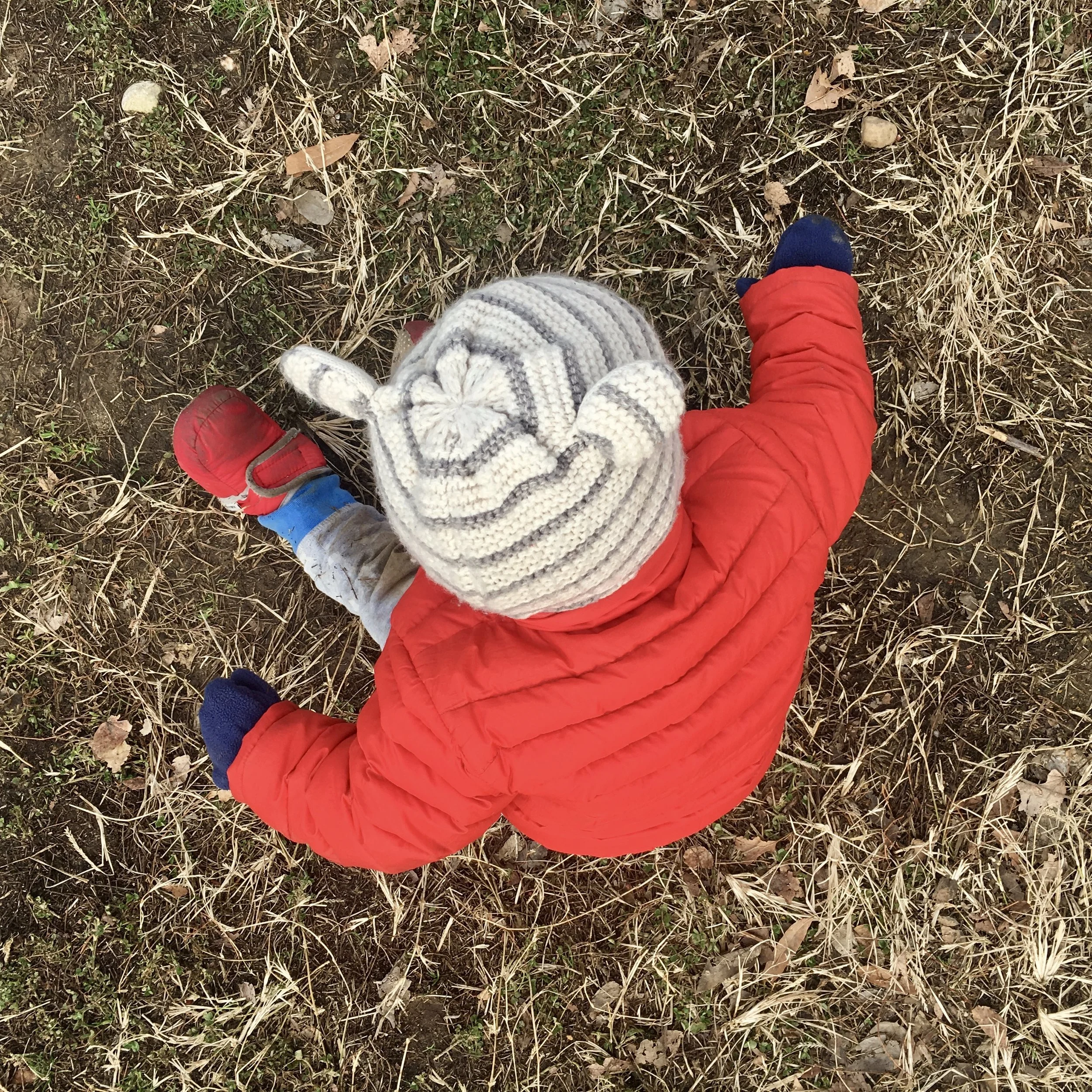 Image: a baby in winter clothes sits in the dirt and dead grass.