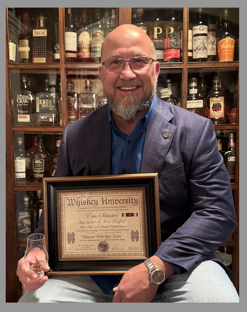 A man with glasses and a beard smiling, wearing a suit jacket and blue shirt, holding a framed certificate that reads "Whiskey University" and a small glass of whiskey, standing in front of a wooden cabinet filled with whiskey bottles.
