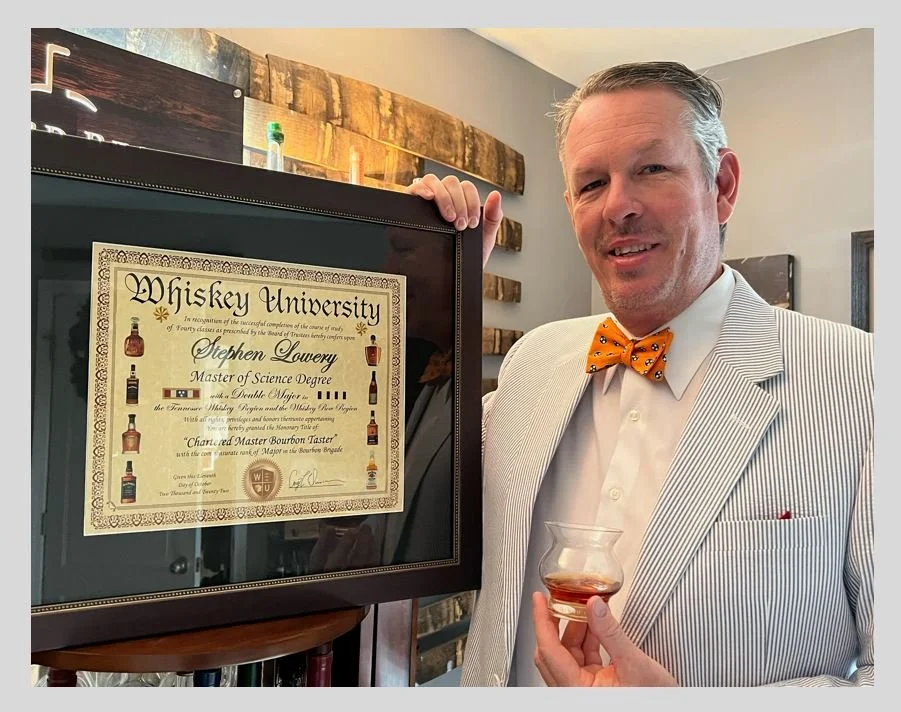 A man in a striped suit with an orange bow tie holding a glass of whiskey and standing next to a framed whiskey university certificate.