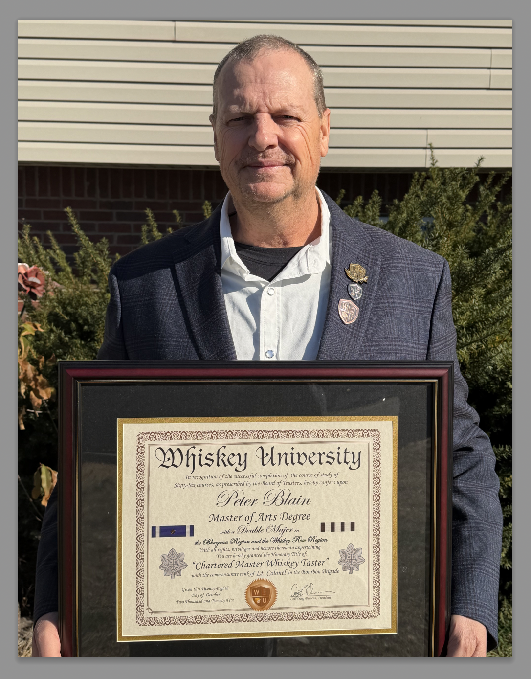 A man in a suit holding a framed diploma from Whiskey University celebrating his graduation with a Master of Arts Degree and Double Major in Charteered Master Whiskey Taster.