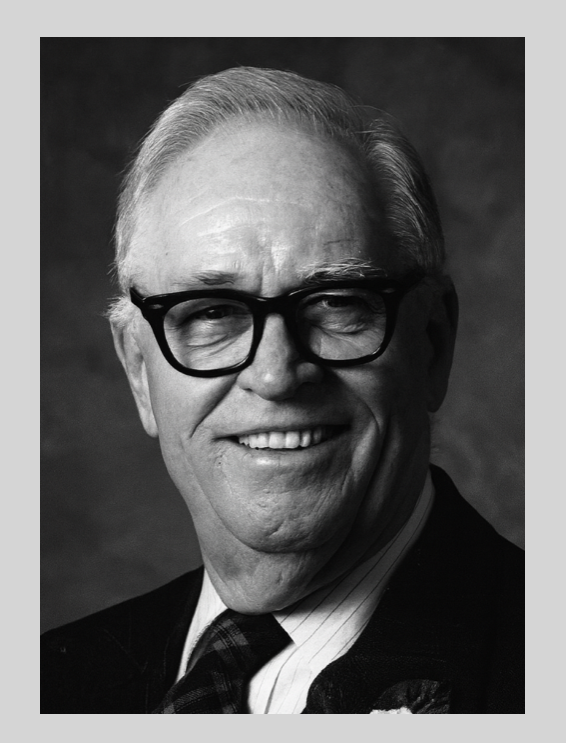 Black and white portrait of an older man wearing thick-rimmed glasses, smiling, dressed in a suit and tie.