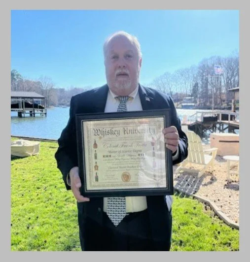 A man wearing a suit holding a framed Certificate from Whiskey University outdoors near a lake