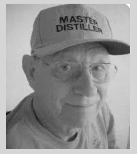 A man wearing a baseball cap with 'Master Distiller' written on it, glasses, and a t-shirt, smiling and taking a selfie.