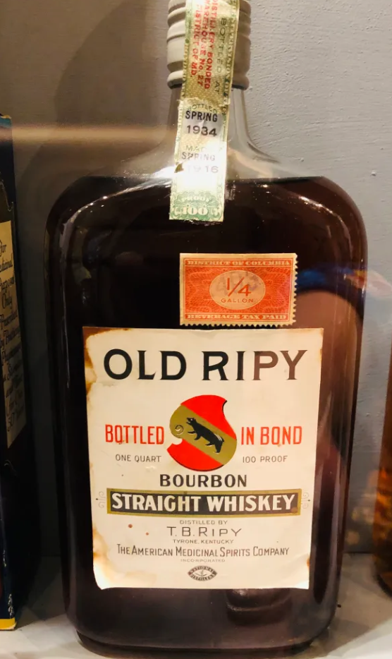 A vintage bottle of Old Ripy bourbon straight whiskey with a large white label, a red and black logo of a lizard, and text indicating it is bottled in bond, 100 proof, and one quart size.