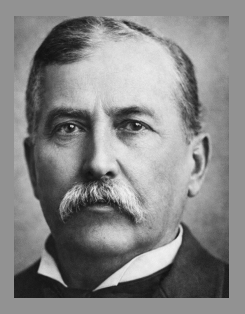 Black and white portrait of a man with a mustache and short hair, wearing a suit and tie.